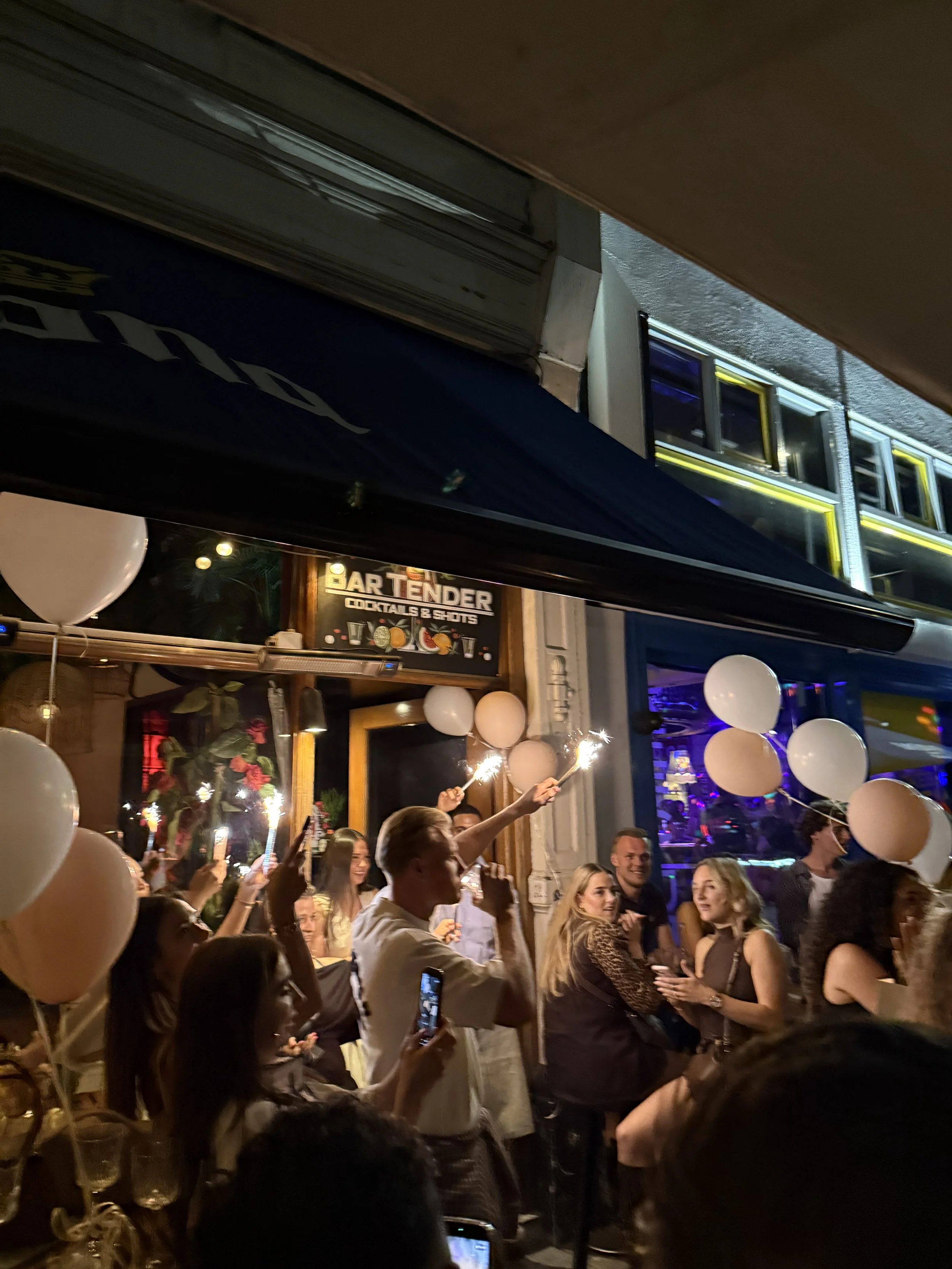 Mensen vieren feest met ballonnen en spuiterijen buiten een café of bar, met een neonbord dat 'Bar Tender' aangeeft, op een avond of nacht.