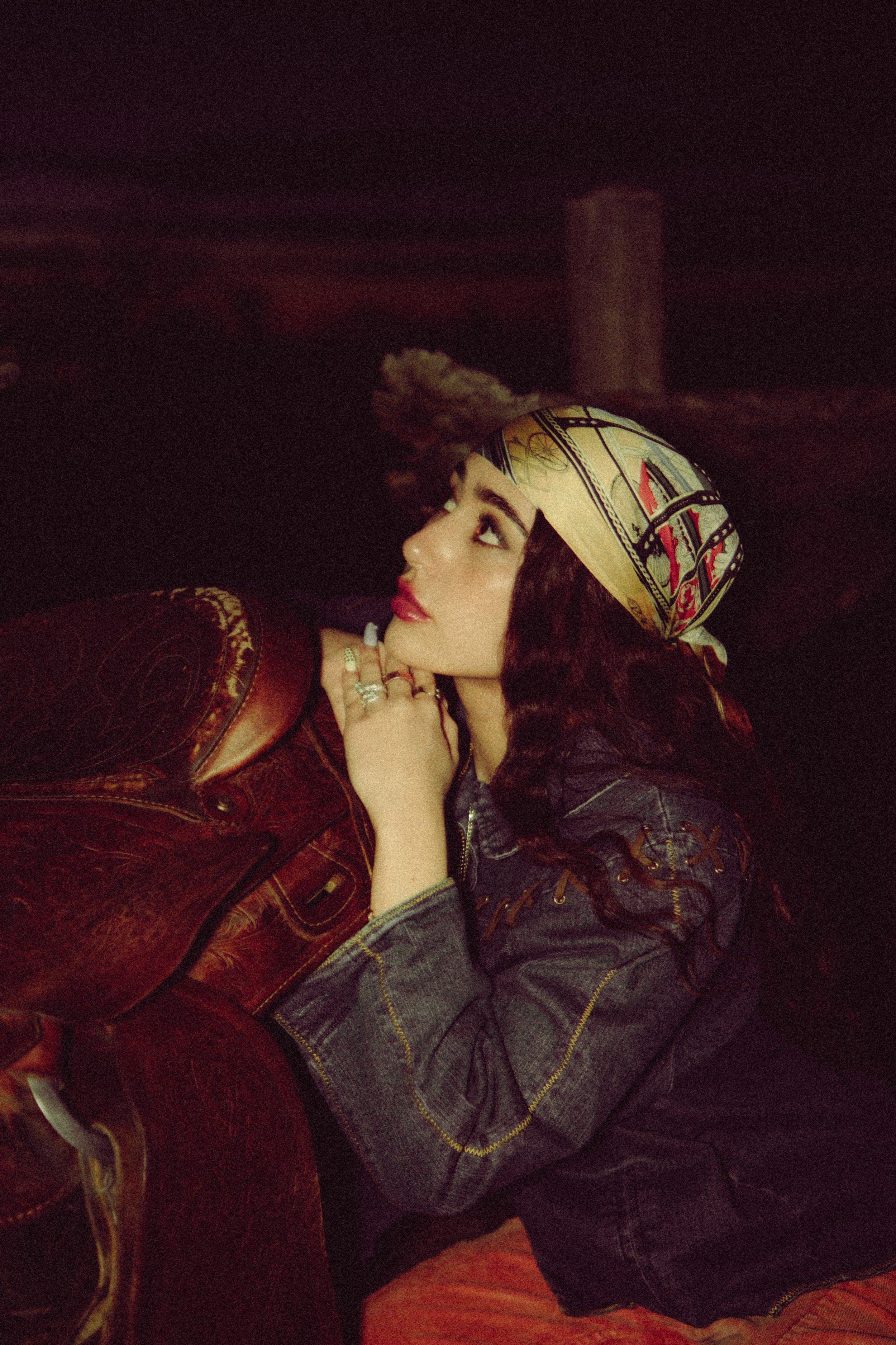 A woman with long curly dark hair, wearing a patterned headscarf, red lipstick, a denim jacket, and rings on her fingers, is leaning her head on a vintage brown leather armchair in a dimly lit setting.