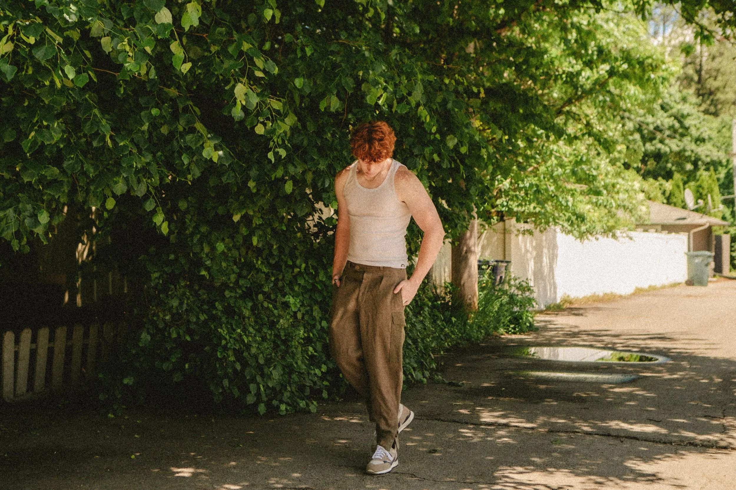 Lifestyle portrait photography by Katsphilm in Albuquerque, New Mexico A person with short curly red hair wearing a beige sleeveless top, brown pants, and sneakers walking along a shaded sidewalk next to green bushes and trees in a residential area.