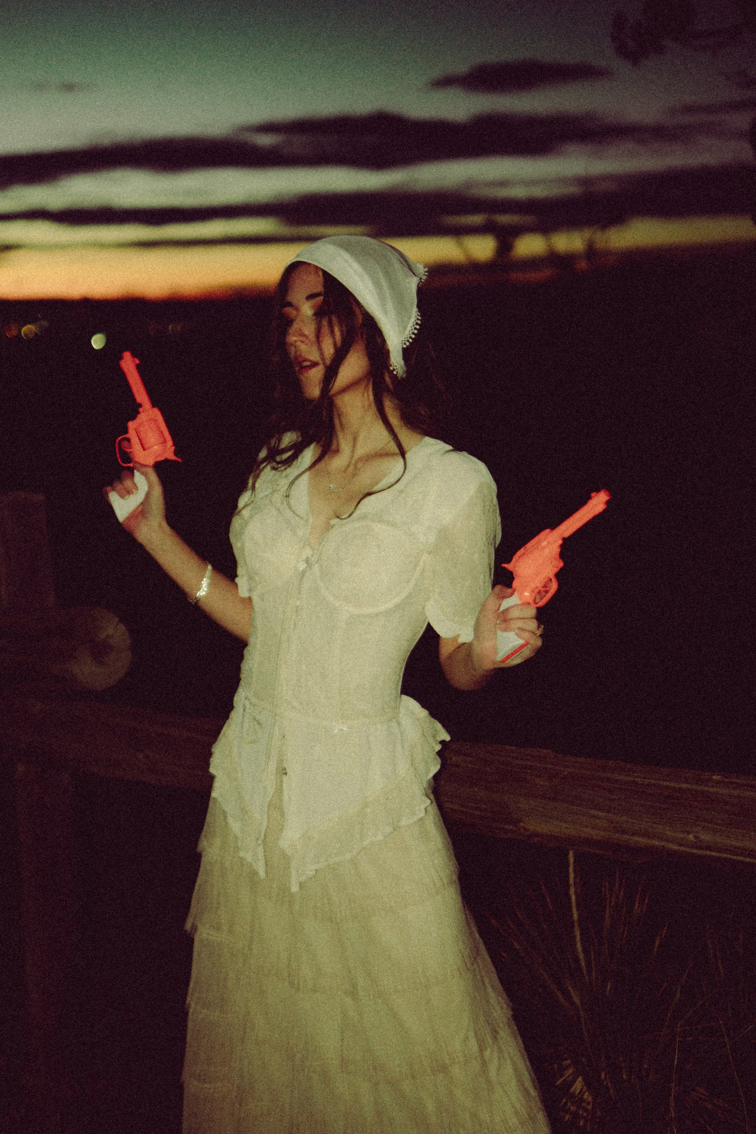 Woman in vintage dress and bonnet holding toy guns at sunset.