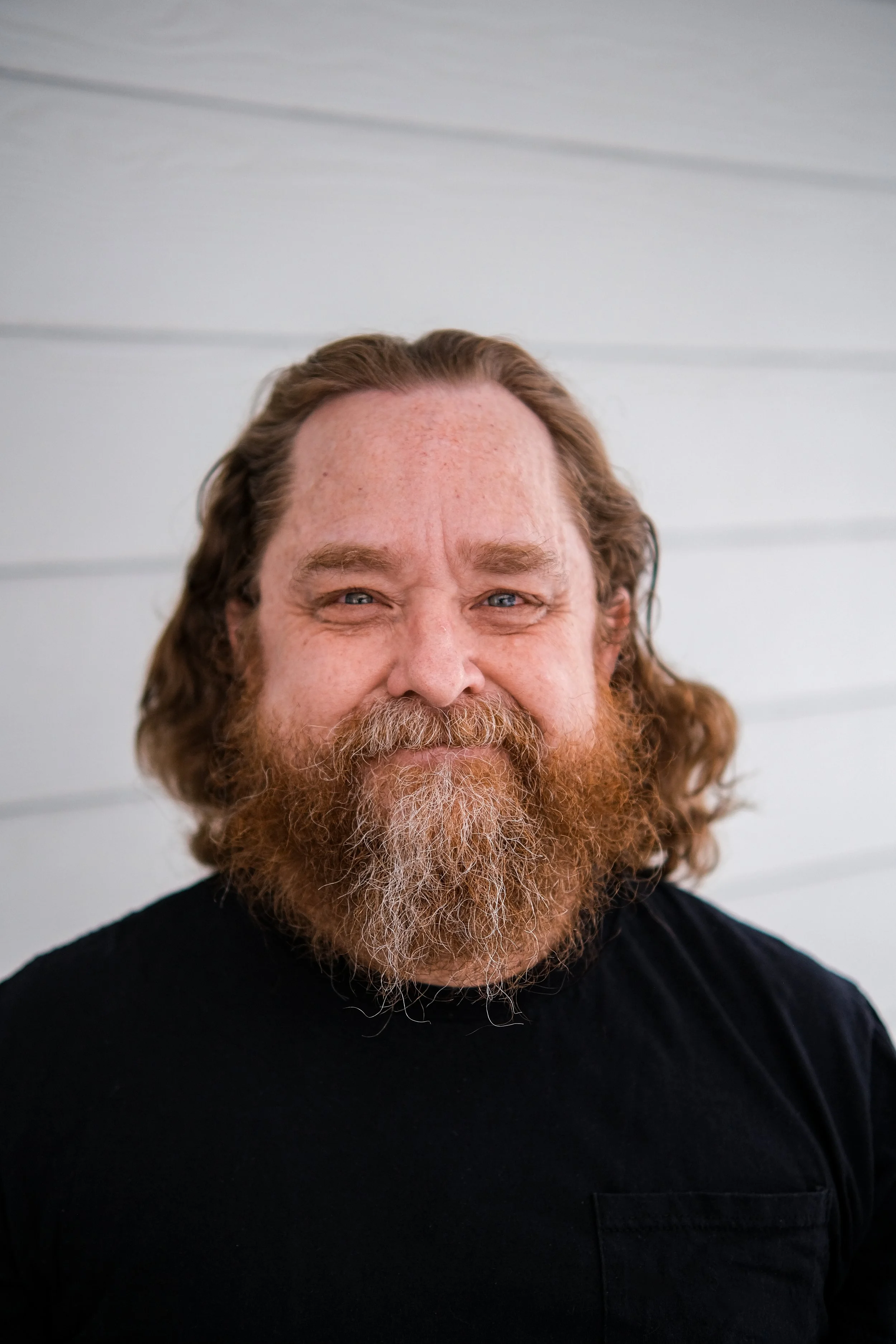 Actor and model headshot photographed by Katsphilm in New Mexico. A man with long hair and a full, reddish beard smiling in front of a light-colored horizontal wood-paneled wall.