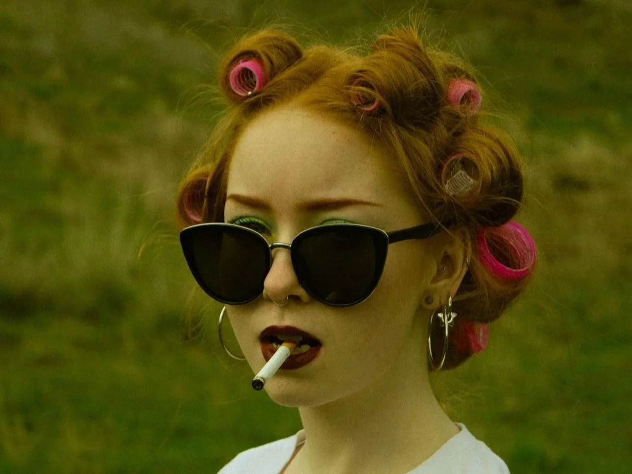 A young woman with red hair in curlers, wearing large sunglasses, hoop earrings, and dark lipstick, smoking a cigarette outdoors.