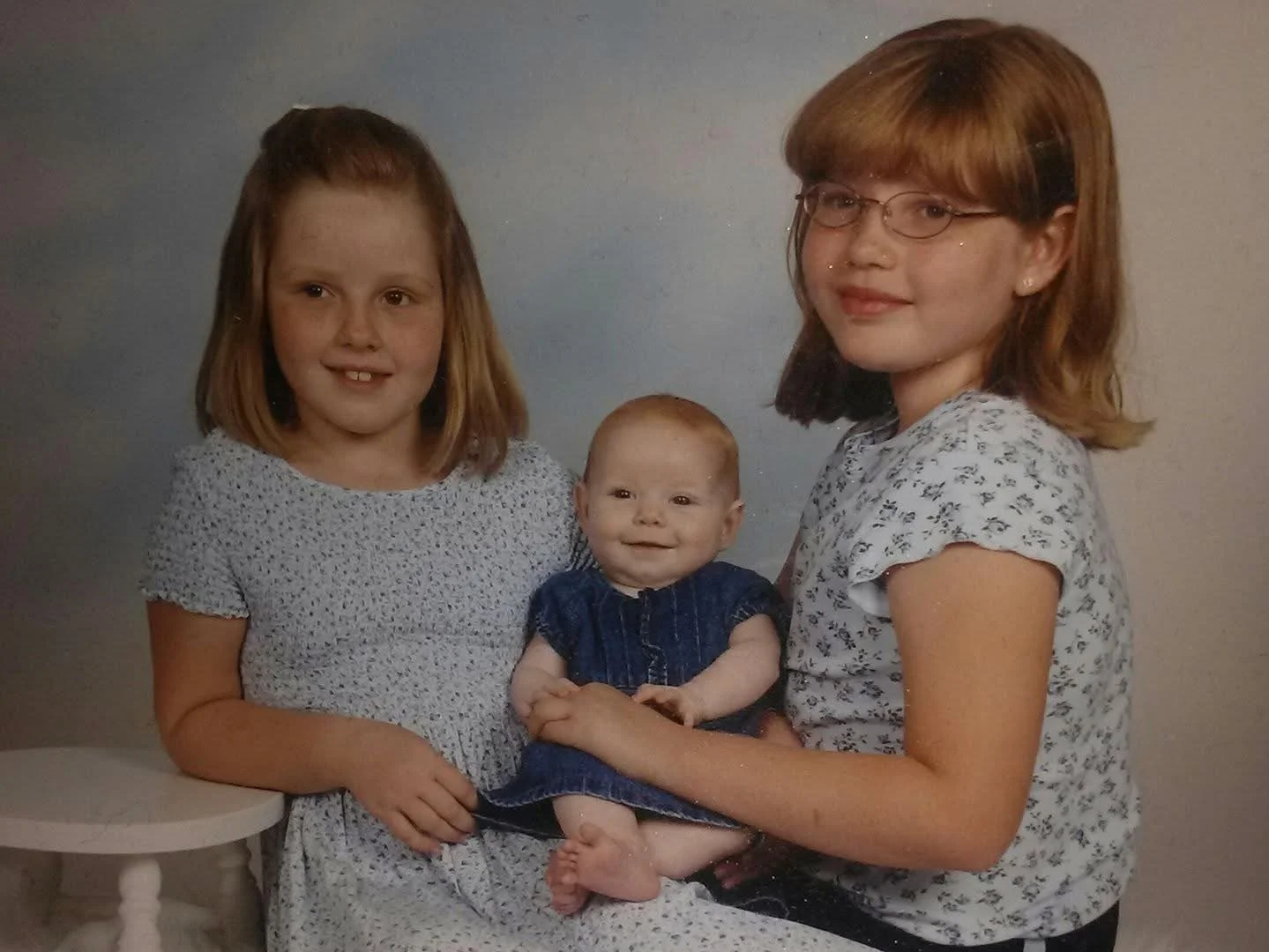 Three young girls, one infant, posing together in a studio setting, all smiling and dressed in casual clothing.