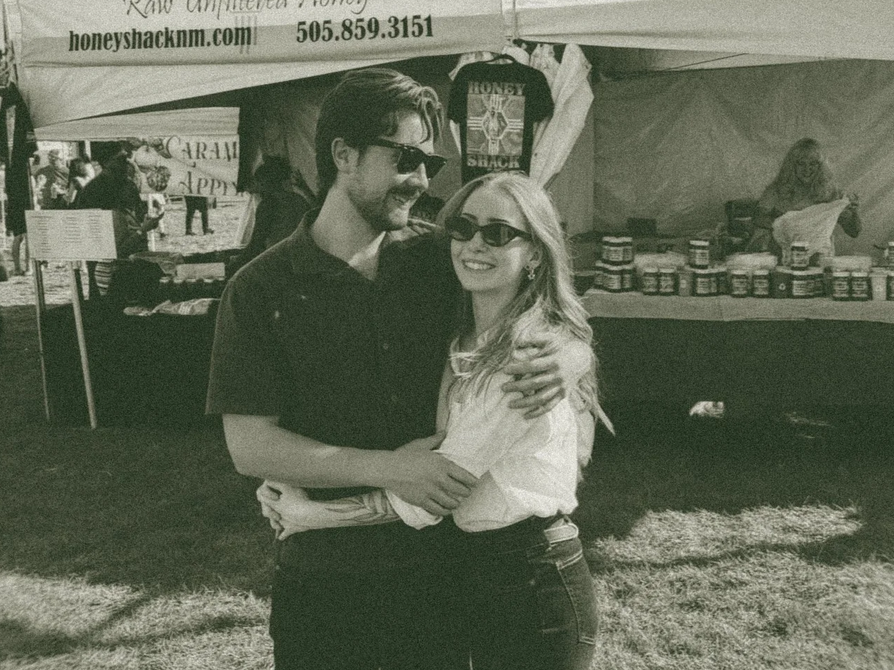 Two young adults, a man and a woman, smiling and hugging at an outdoor event or fair, both wearing sunglasses. The background shows a booth with jars, a woman behind the booth, and tents.