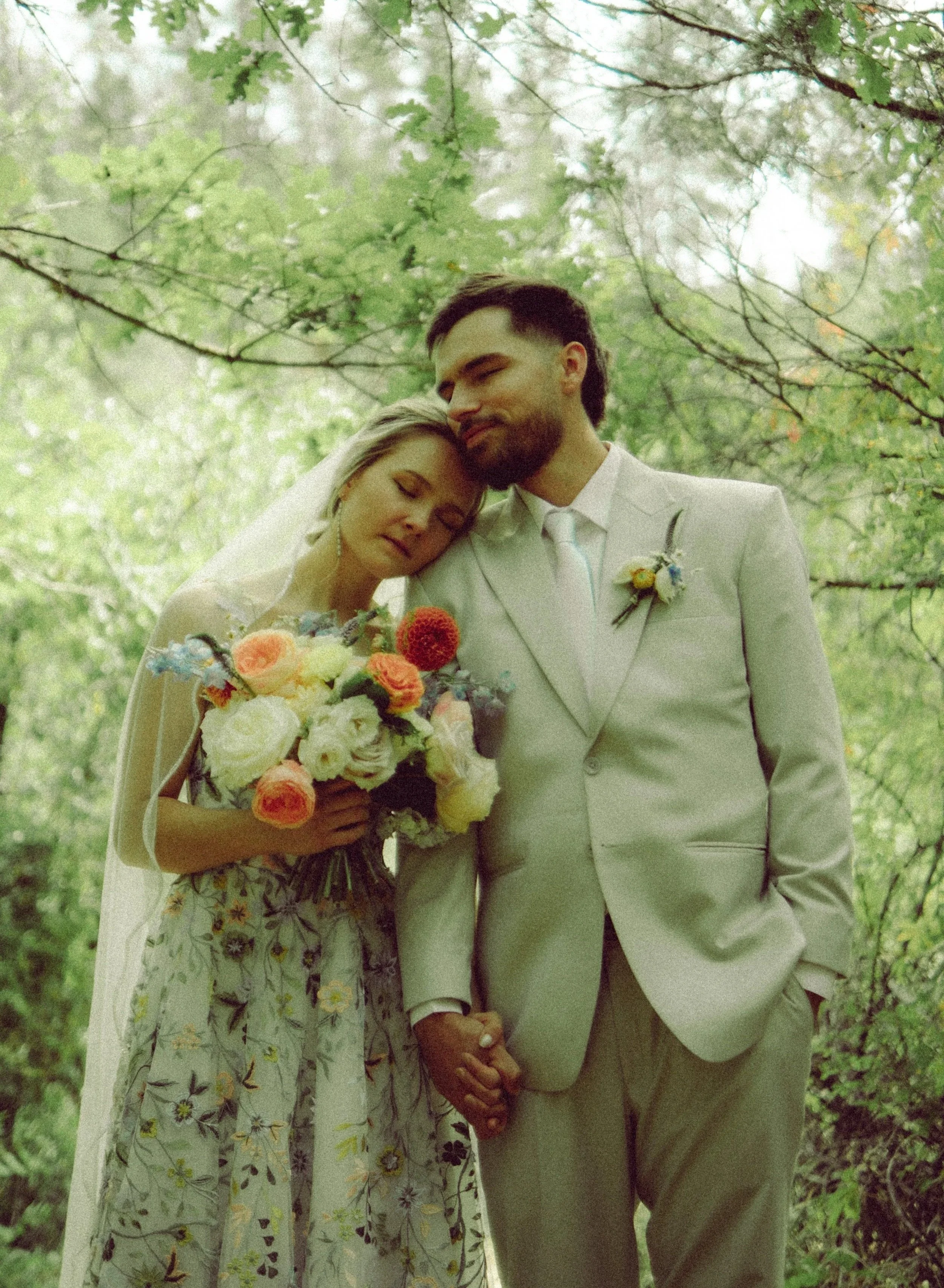 A bride and groom in wedding attire stand close together outdoors in a forest, with the bride resting her head on the groom's shoulder, both holding hands; the bride holds a bouquet of colorful flowers.