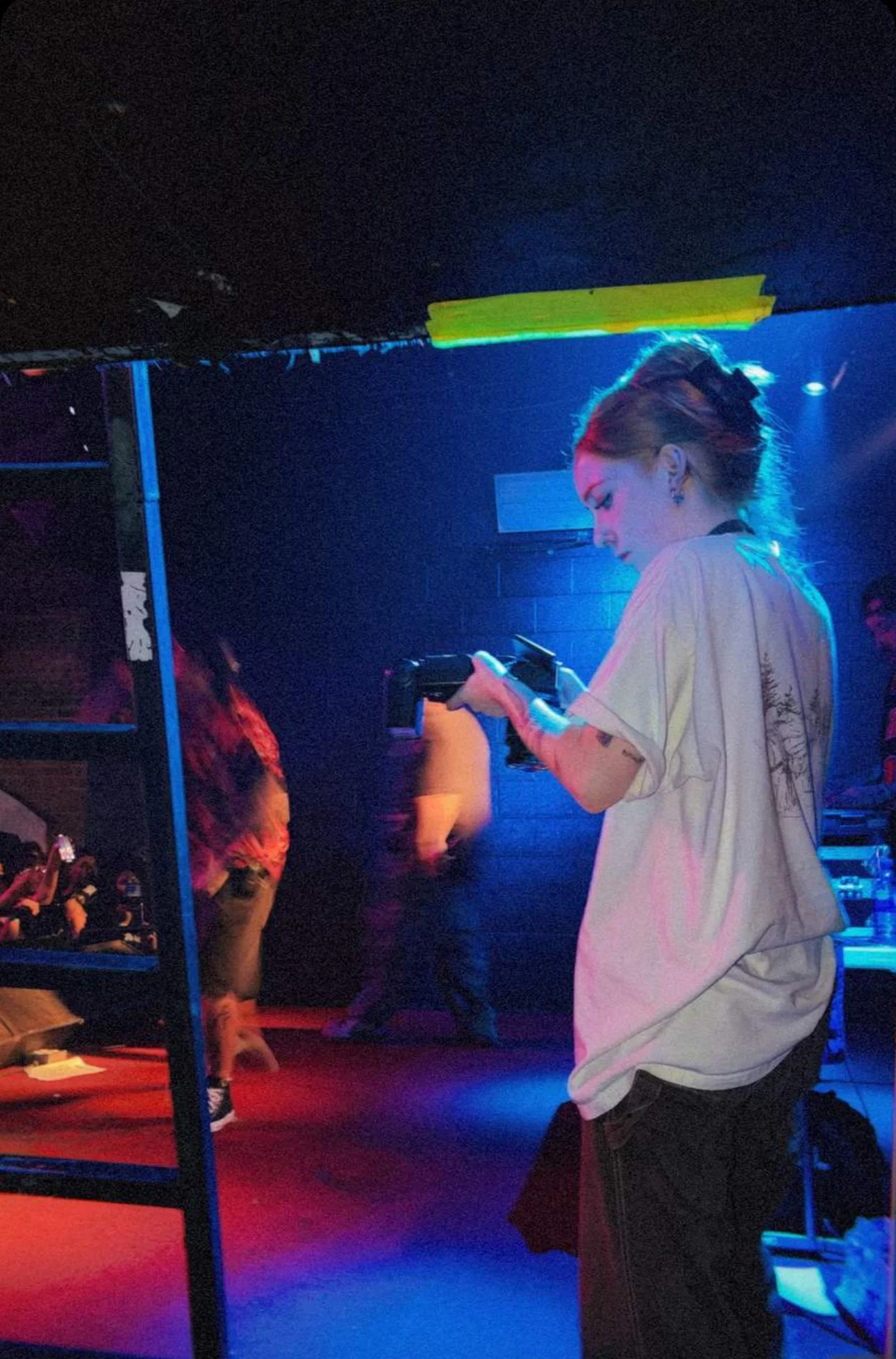 A woman holding a camera in a dark, colorful, club-like environment, with people in the background and neon lights.