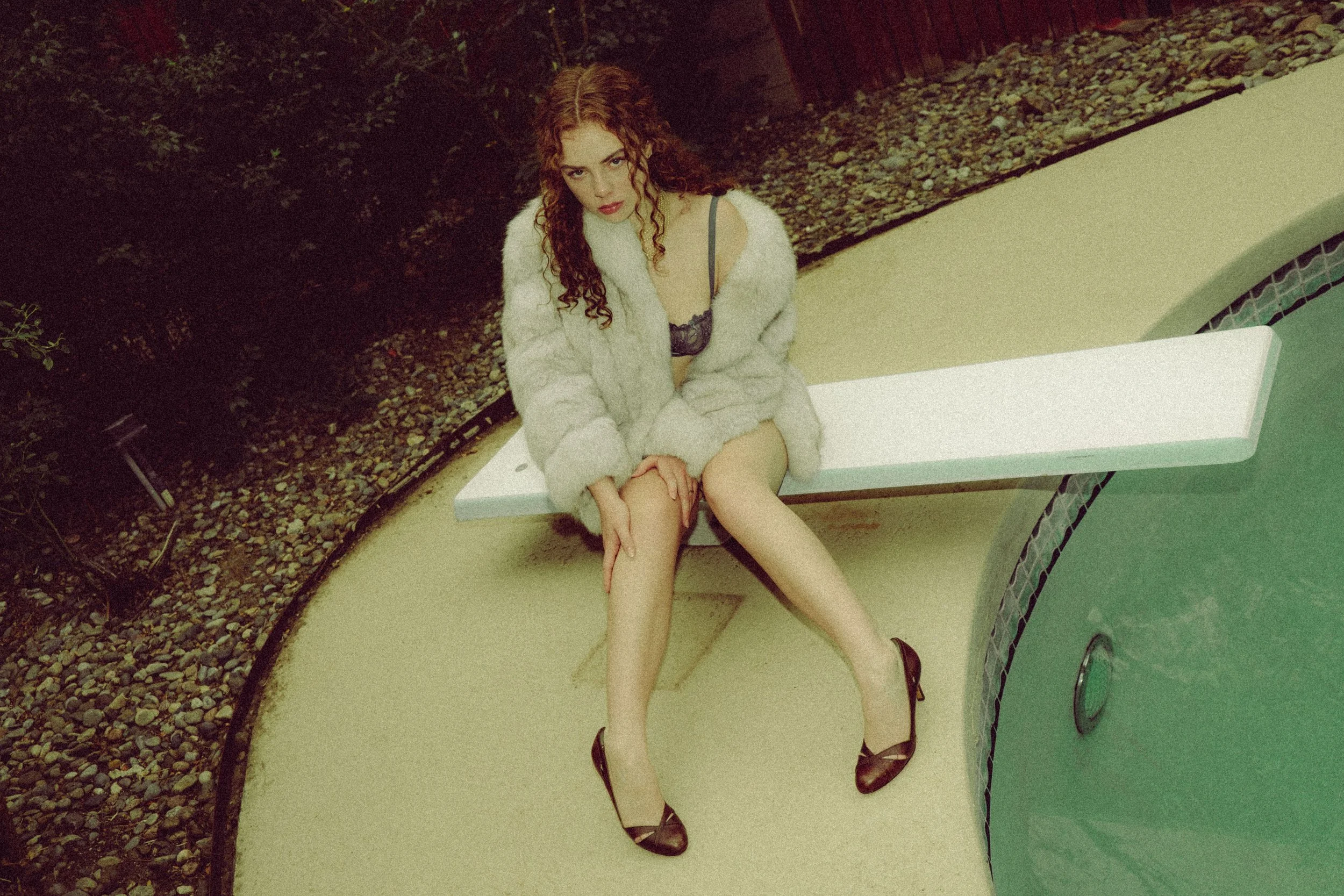 Creative personal branding portrait of a woman photographed by Katsphilm. A woman with curly red hair sitting on a white bench near a pool, wearing a black bra, a white fur coat, beige stockings, and black heels.