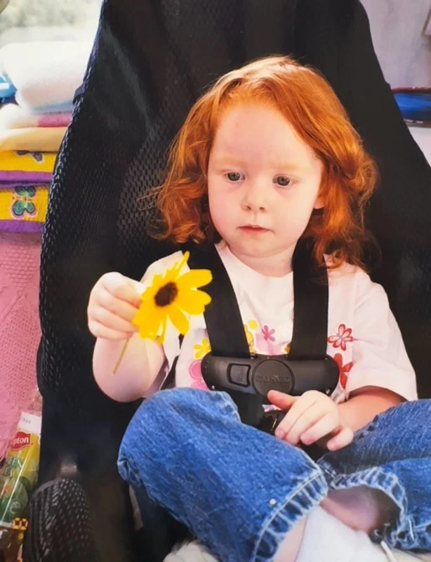 A young girl with red hair sitting in a stroller, holding a yellow flower with a dark center, looking intently at it.