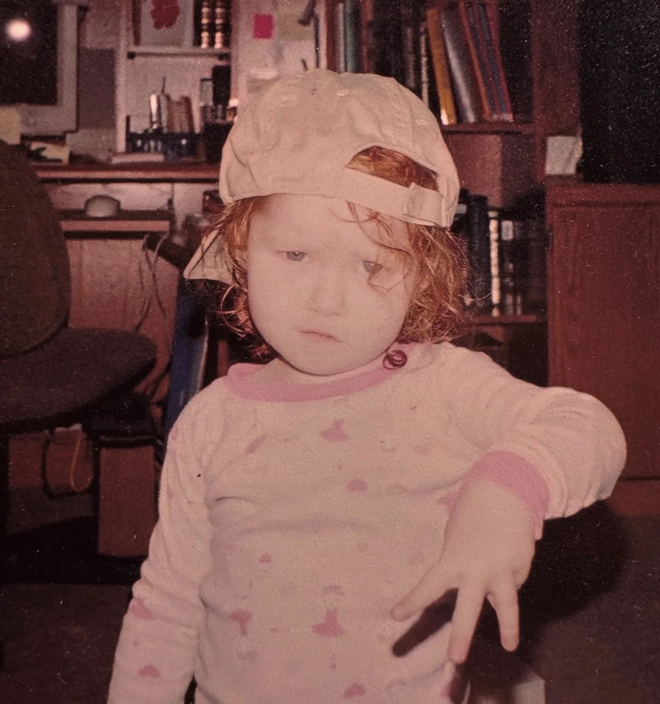 A young girl with curly red hair wearing a pink long-sleeve pajama top with a rainbow pattern and a baseball cap backwards, in a room with shelves of books and furniture in the background, reaching out with her fingers.