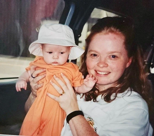 A woman holding a baby inside a vehicle, with the woman smiling and the baby wearing an orange dress and a white sun hat.