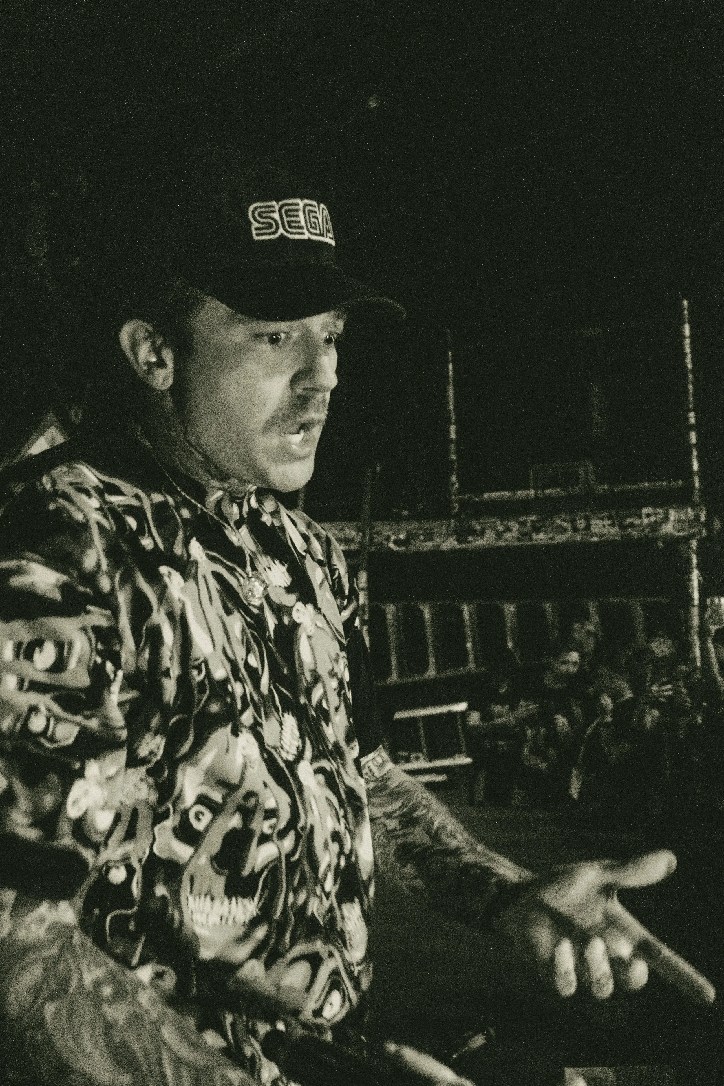 A rapper named Terror Reid standing outdoors at night, wearing a cap with 'SEGA' logo, a patterned shirt, and displaying tattoos on his arms, gesturing with his hand.