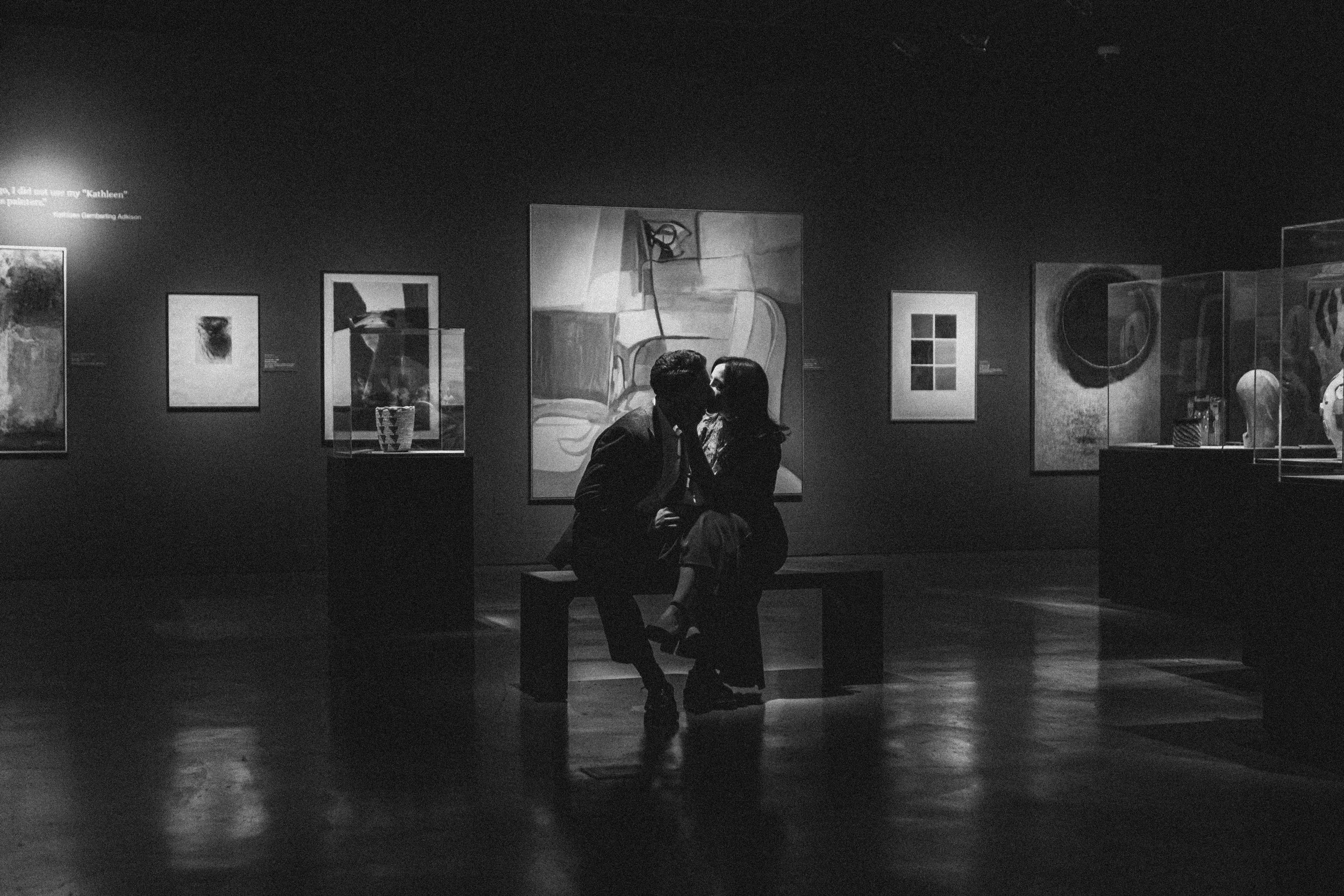 Silhouetted couple sitting on a bench in an art gallery, sharing a kiss amidst various paintings and sculptures on display.