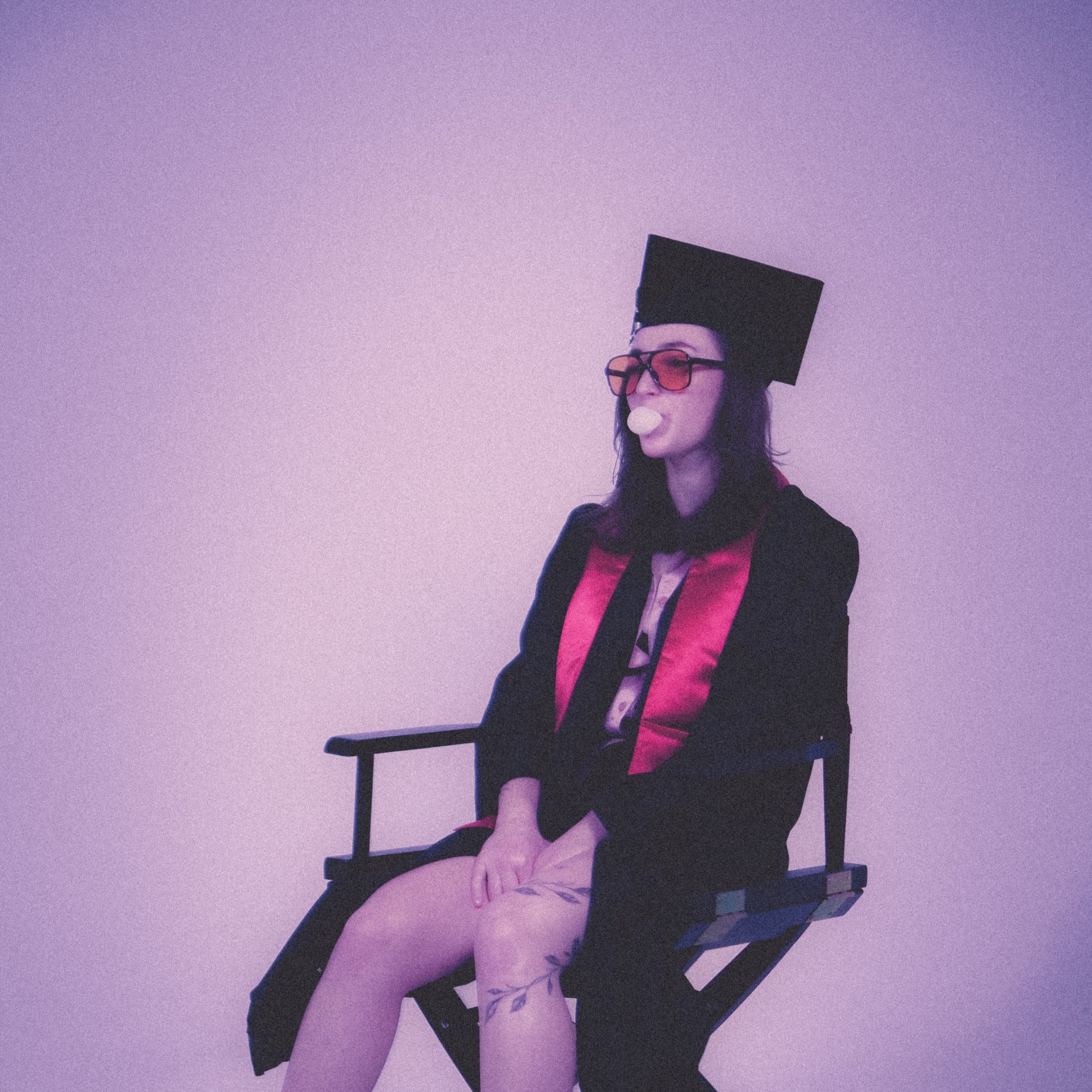 A woman in academic regalia, including a cap and gown, sitting on a director's chair against a plain wall.