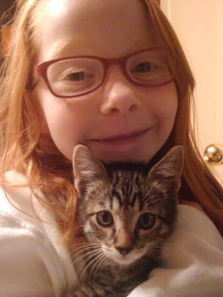 A young girl with red hair, glasses, and a white shirt holding a tabby kitten with striped fur.