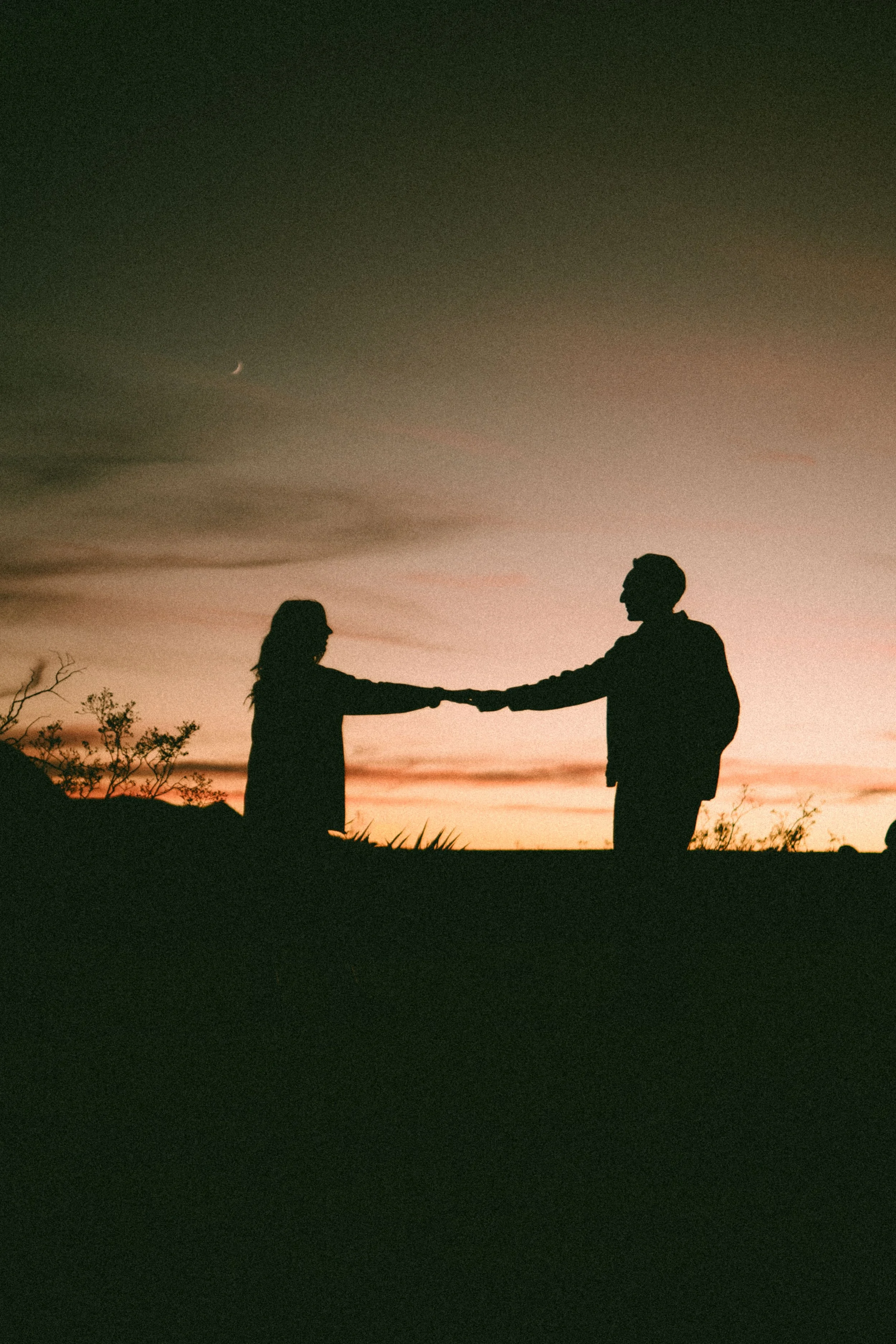 Couple holding hands at sunset during an engagement session in New Mexico. Silhouette of a man and woman holding hands at sunset with a crescent moon in the sky.