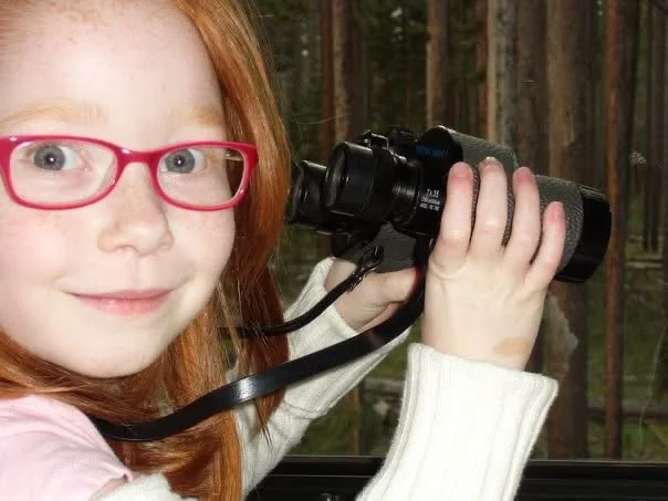 A young girl with red hair and red glasses looking out a window while holding a black telescope. The background shows a forest with tall trees.