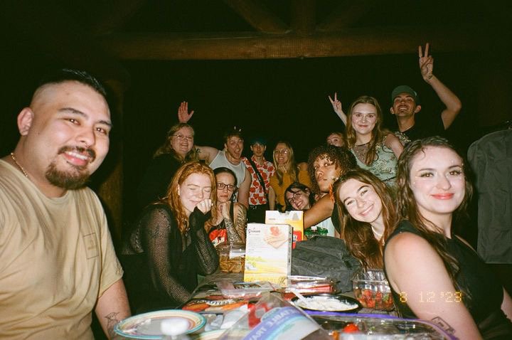Group of young adults sitting at a table in a dimly lit restaurant or bar, smiling and making peace signs, with some raising their hands in the background.
