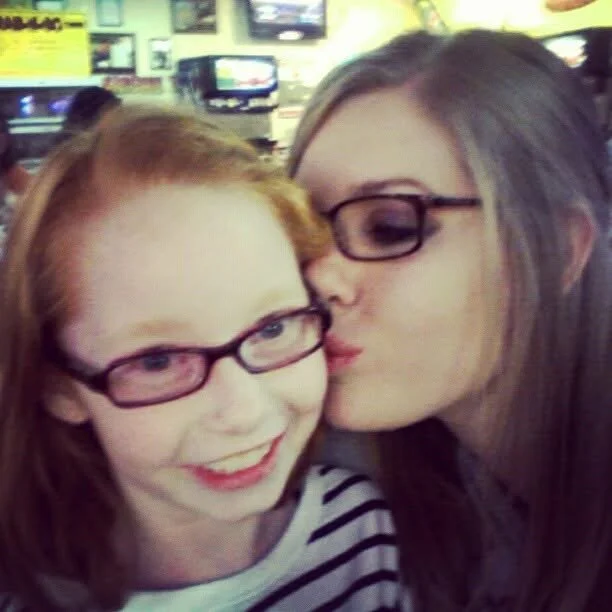 Two women with glasses sharing a kiss on the cheek at a restaurant or cafe.