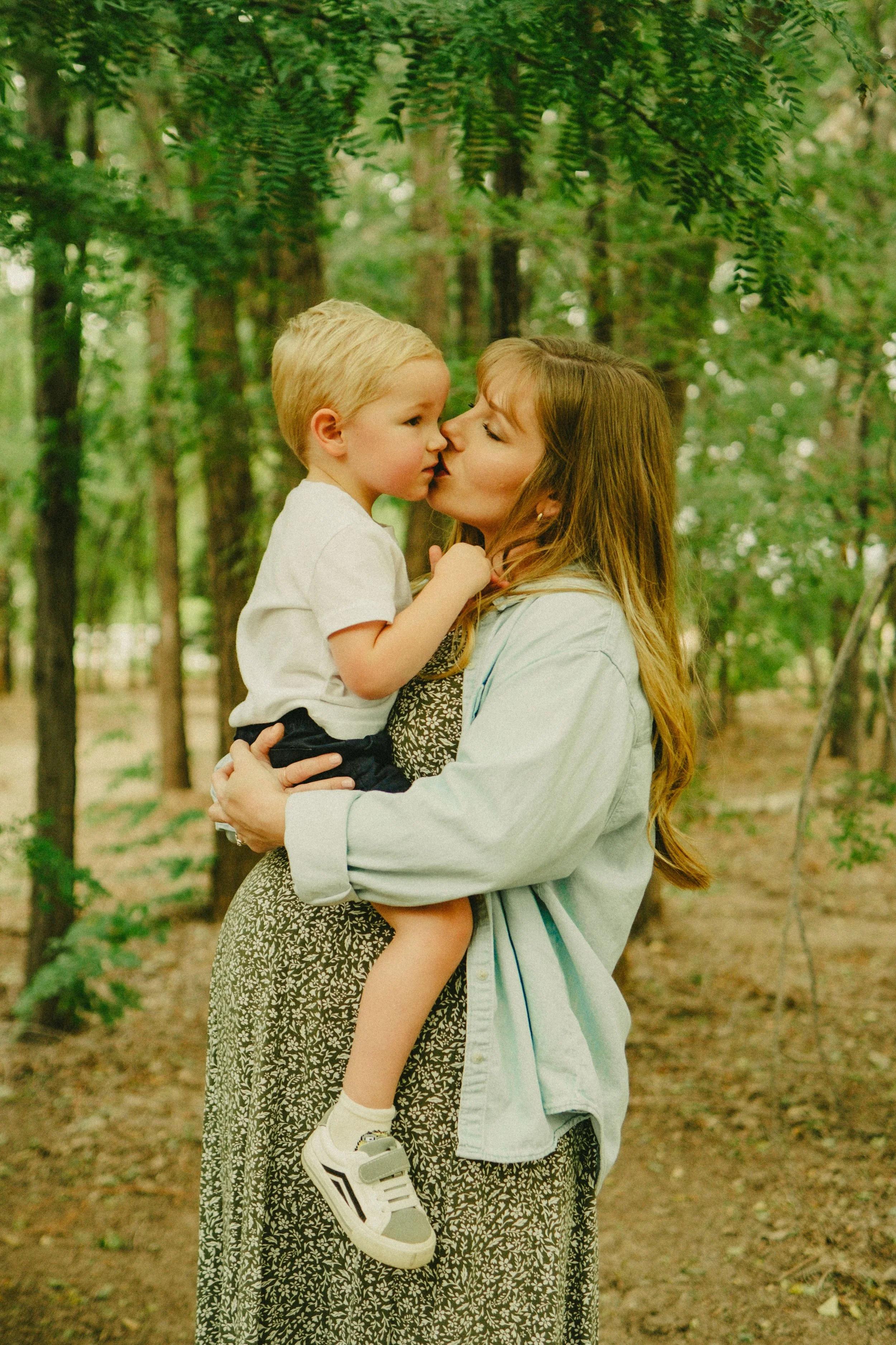 A woman with long, light brown hair holding a young blonde boy in a forest. The woman is giving a kiss on the nose to the boy, who is sitting on her arm. The scene is surrounded by green trees and foliage.