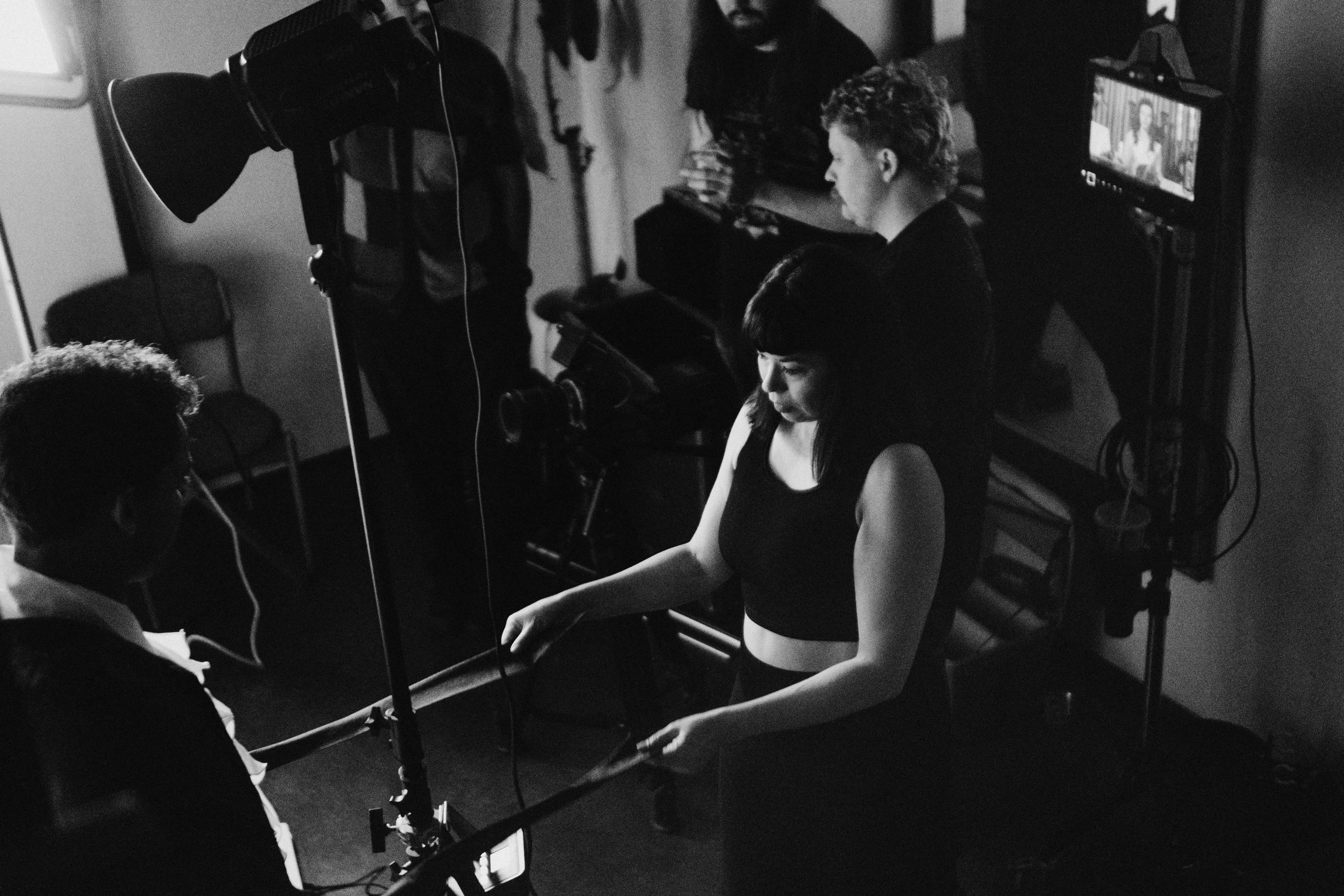 Black and white photo of a film set with a woman in a sleeveless top and a man with curly hair standing in front of a camera. Crew members are working around her with a large light and a monitor on a stand to the right.