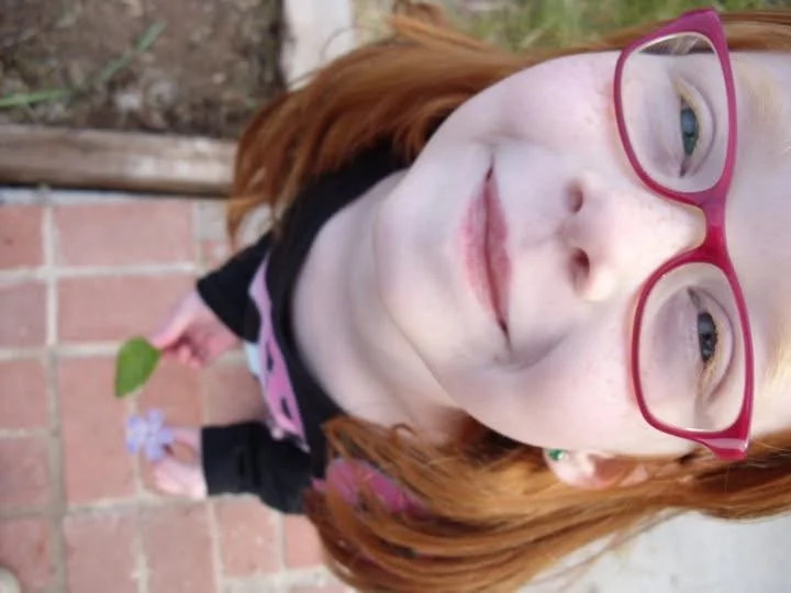 Close-up of a young girl with red hair wearing pink glasses and a black and pink shirt, standing on a brick patio holding a leaf and a small flower in her hand.
