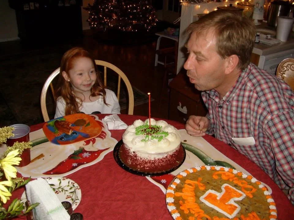 A man and young girl celebrating a birthday at a dinner table with a cake, decorated with a single candle, and a pizza. The girl looks excited, and Christmas decorations are visible in the background.