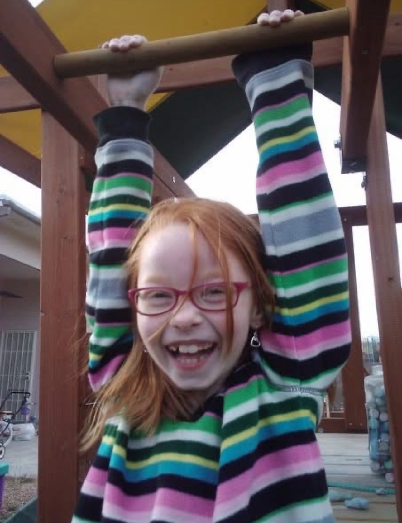 A girl with red hair, wearing glasses and a colorful striped sweater, hanging upside down from a wooden play structure on a playground.