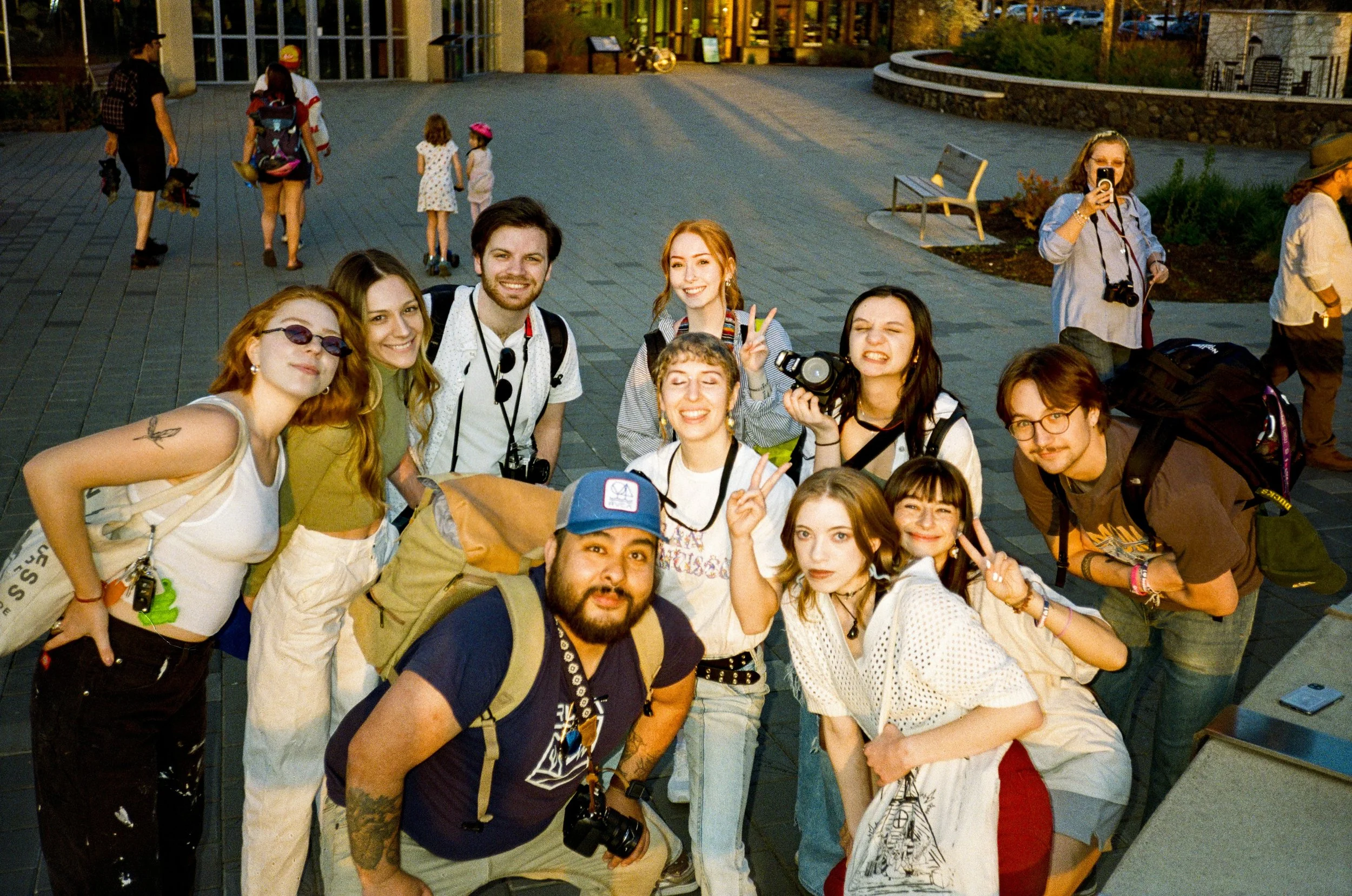 A group of smiling young adults and teenagers posing for a photo outdoors in the evening, some making peace signs, with a cityscape in the background.