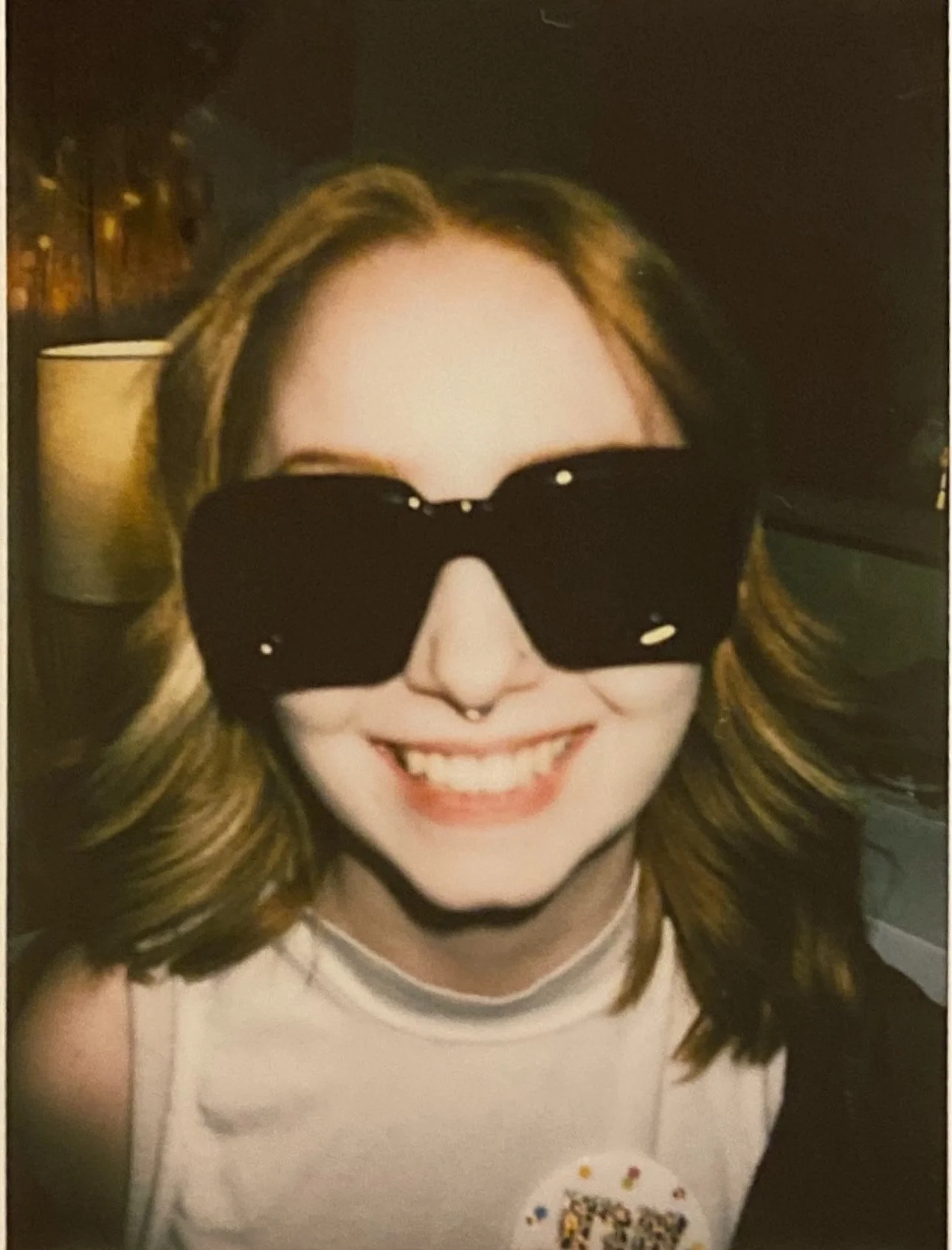 A woman with shoulder-length light brown hair wearing large black sunglasses and a septum piercing, smiling widely. She has a white top with a decorated badge or button on it.