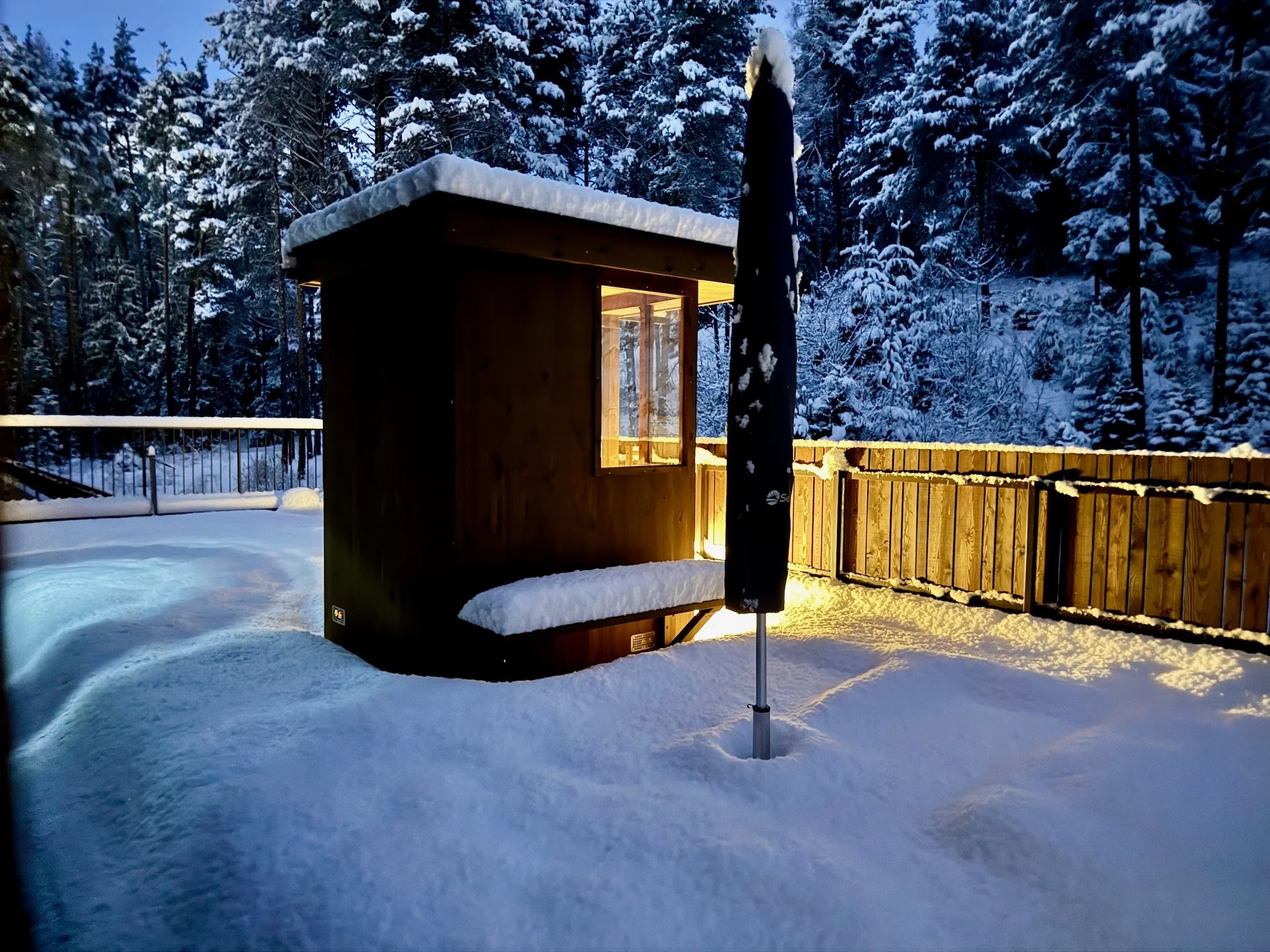 Private Holzsauna im verschneiten Garten mit warm beleuchtetem Fenster, umgeben von Winterwald und Holzzaun.