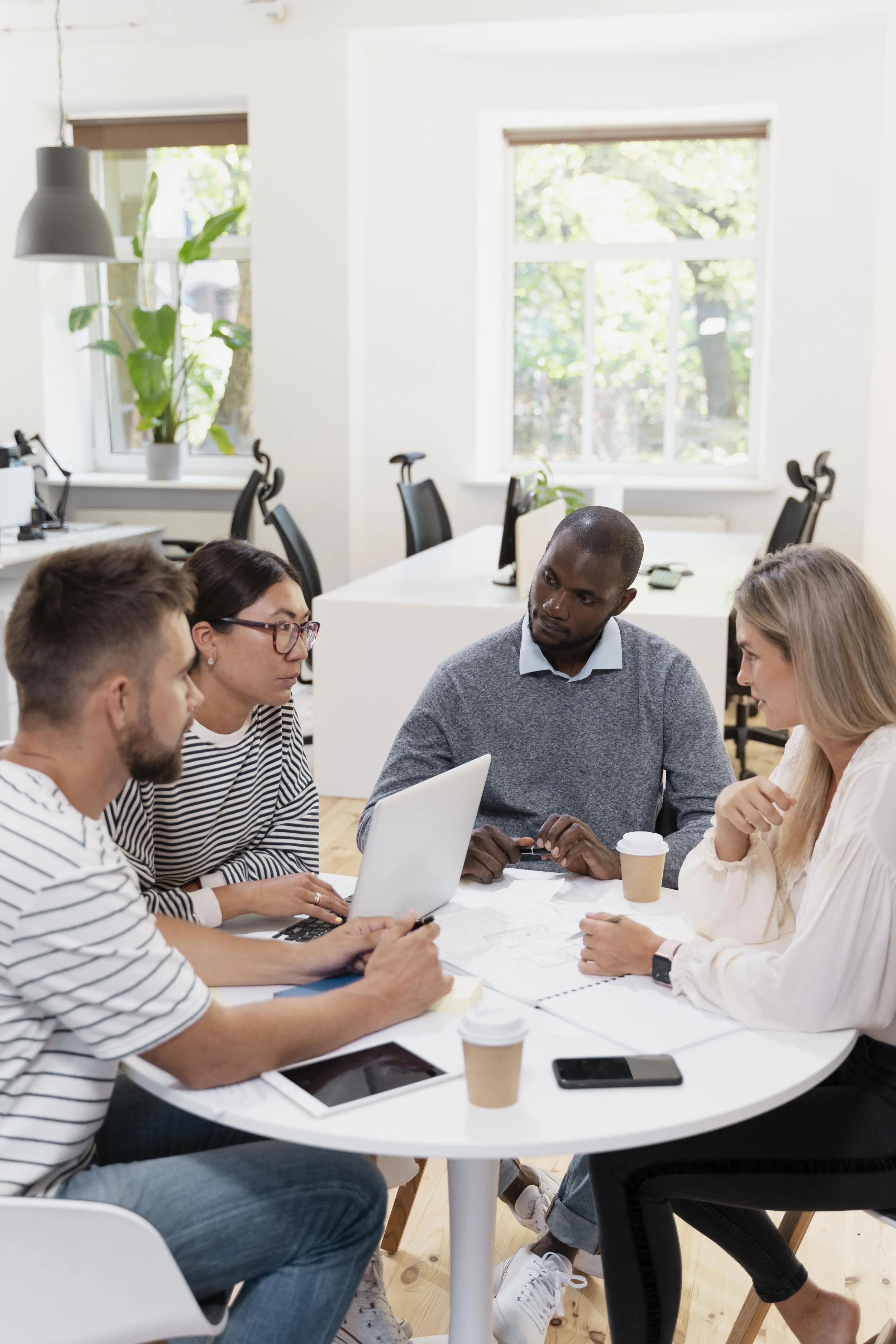 A group of five diverse young professionals engaged in a meeting around a white table in an office, with laptops, notebooks, coffee cups, and a smartphone.