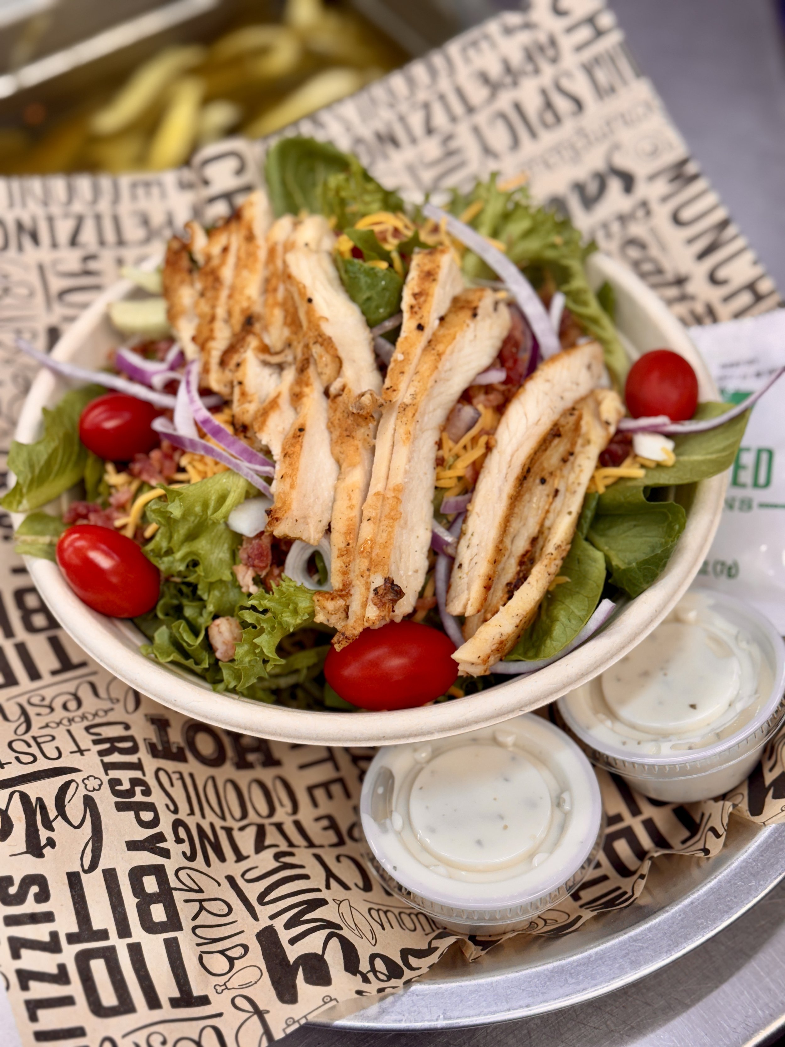 A salad with grilled chicken, cherry tomatoes, shredded cheese, and lettuce in a takeout container, with small containers of ranch dressing on the side.