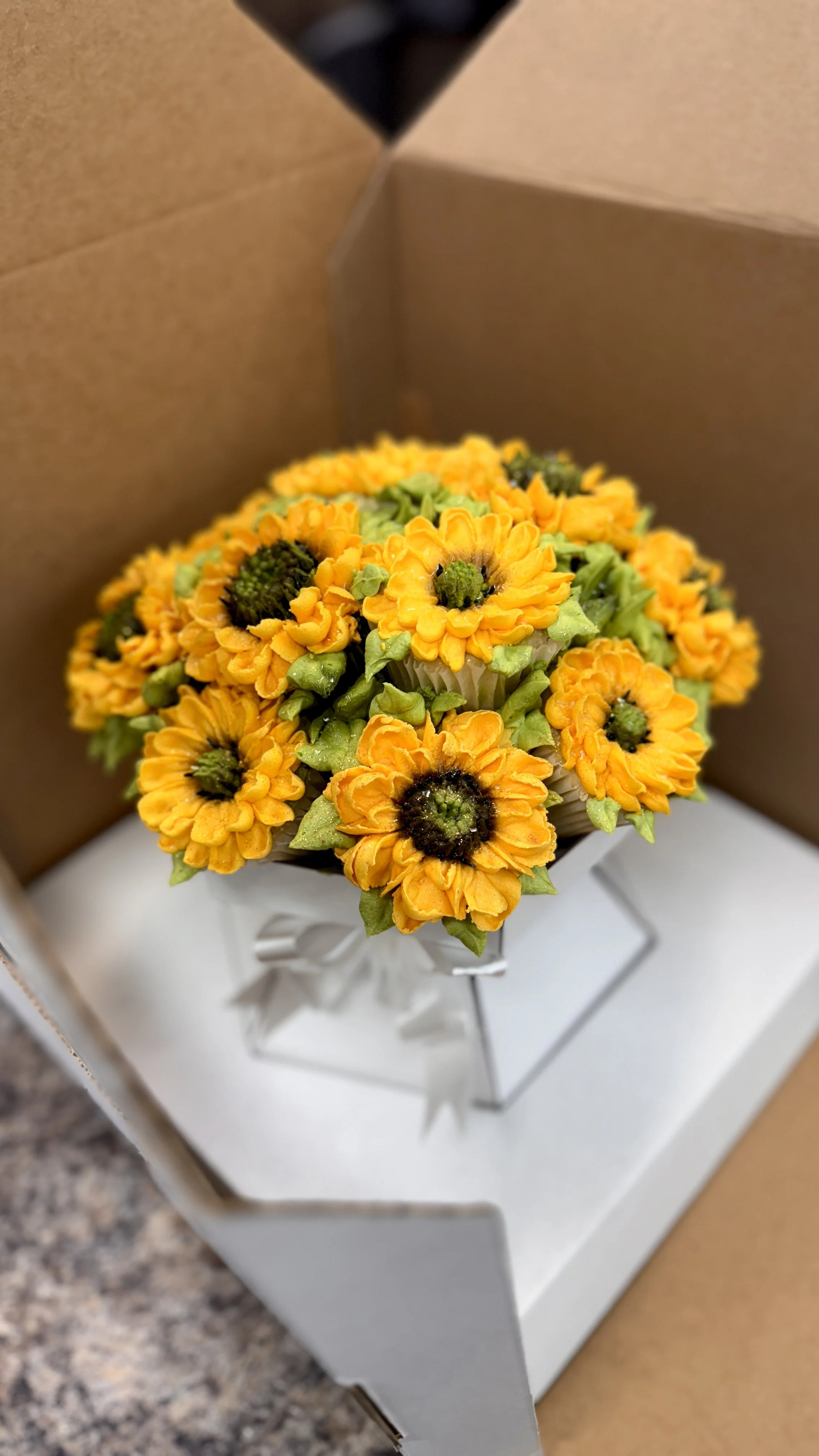 Bouquet of yellow and orange daisy-like flowers with green centers, wrapped in white ribbon, in a white box.