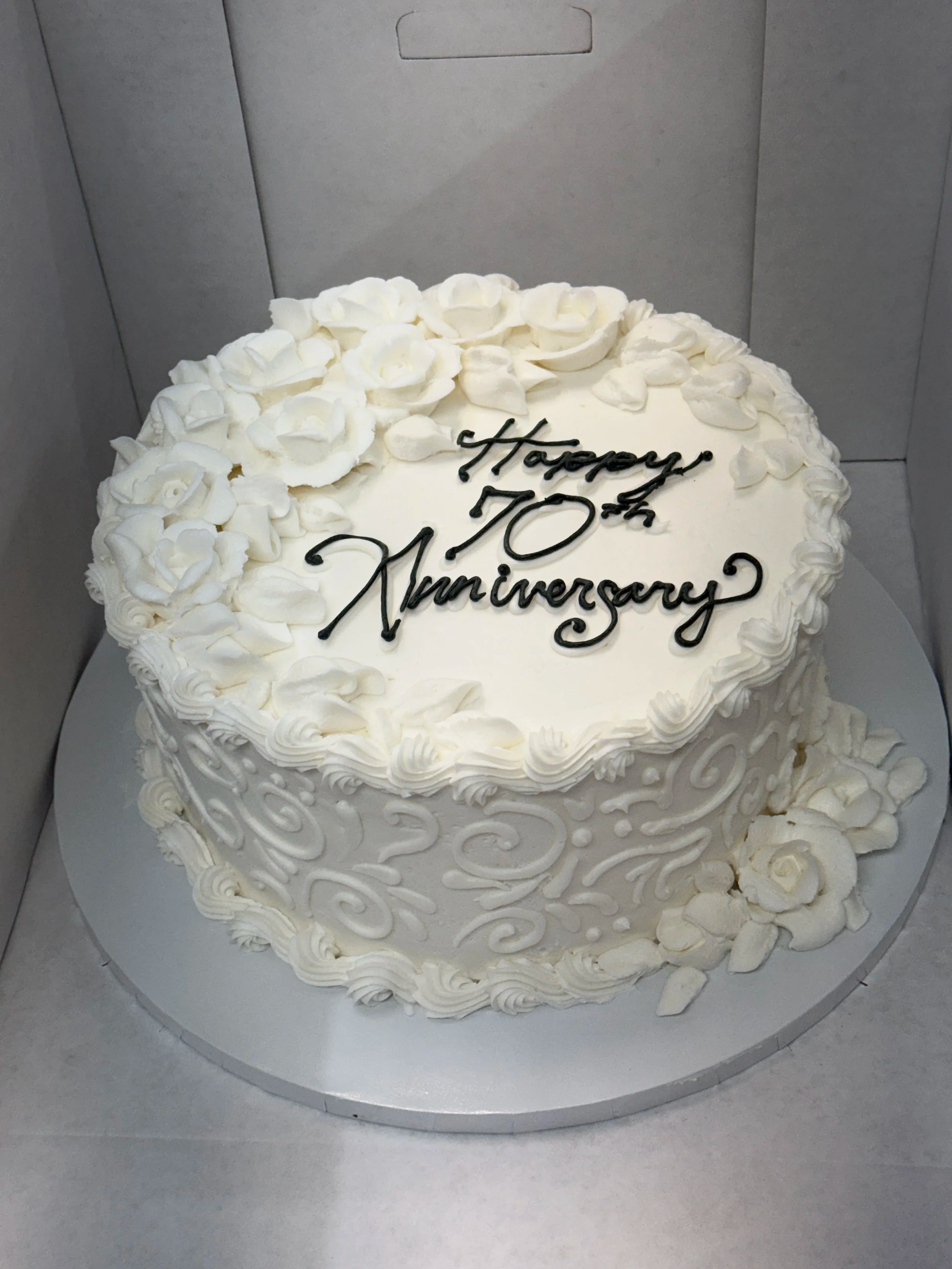 A white, round cake with floral decorations, piping, and a message that reads 'Happy 10th Anniversary' in black icing on top.