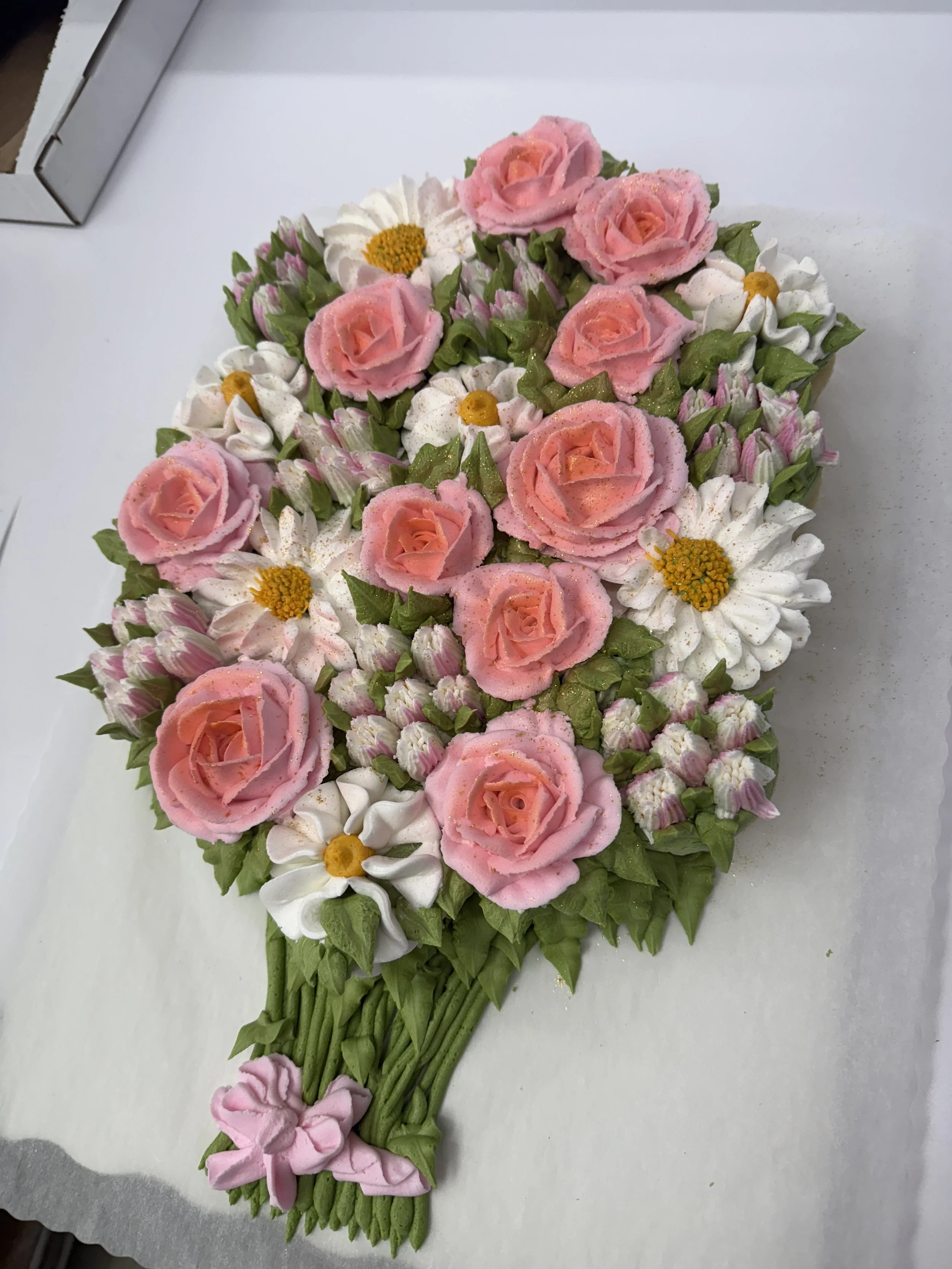 A decorative floral arrangement made of pink roses, white daisies with yellow centers, and green leaves, laid out in a teardrop shape on a white surface.