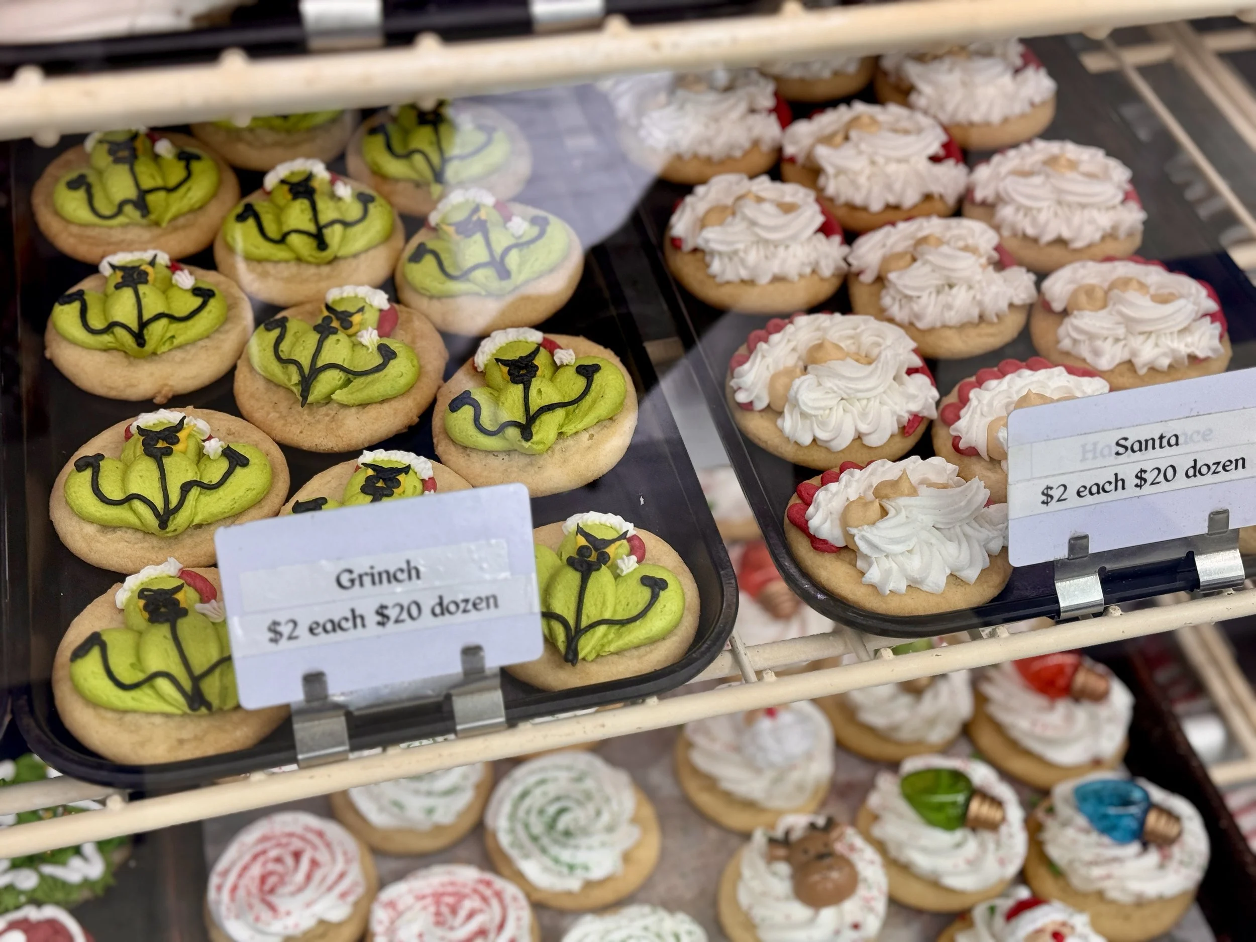 Display case of decorated cookies featuring characters from Dr. Seuss, including the Grinch with green frosting and a red and white frosting cupcake decorated with white icing, red and green accents, and small tan candies.