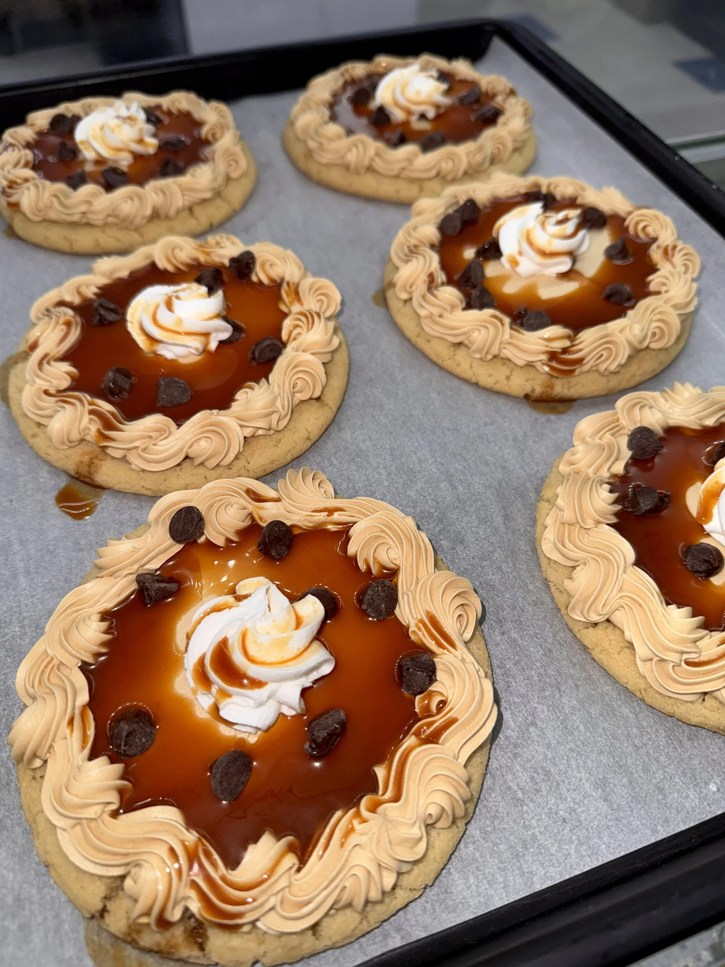 Four heart-shaped cookies with caramel, whipped cream, chocolate chips, and decorative piping on parchment paper.