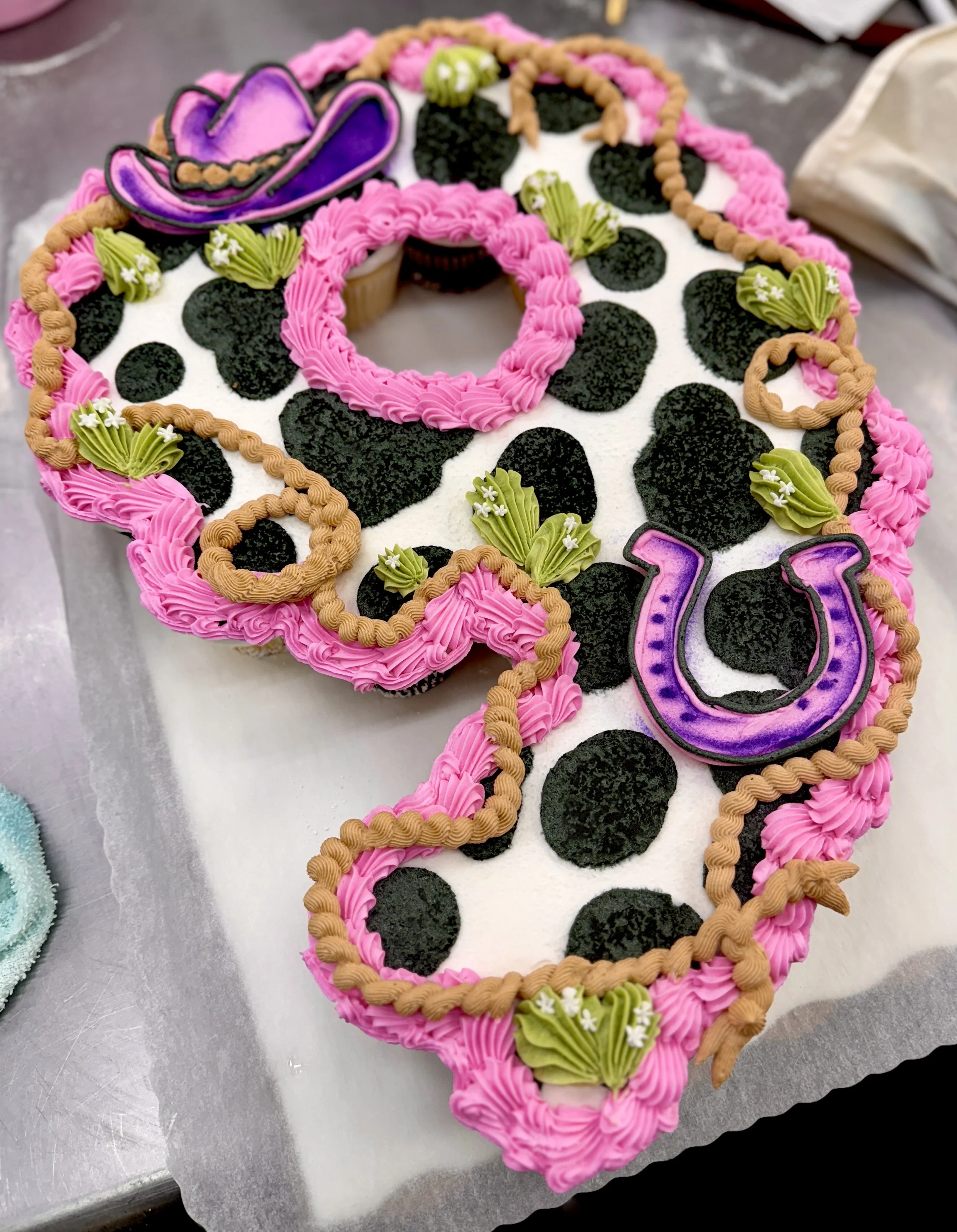 Pink and black decorated cake in the shape of a tequila bottle with cowboy hat, flowers, and geometric designs.