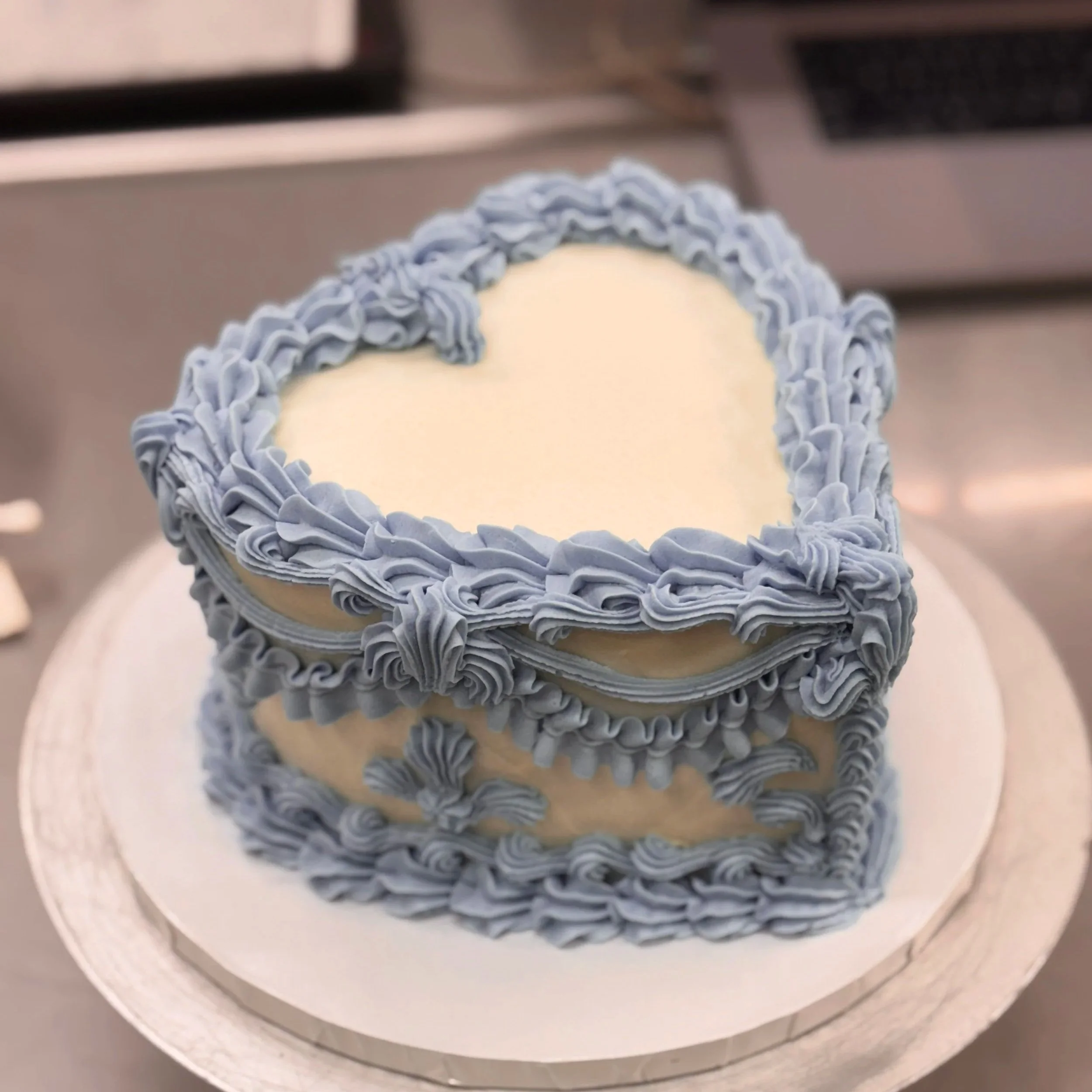 Close-up of a round cake with white frosting, decorated with blue ruffled icing around the edges, placed on a white cake board.