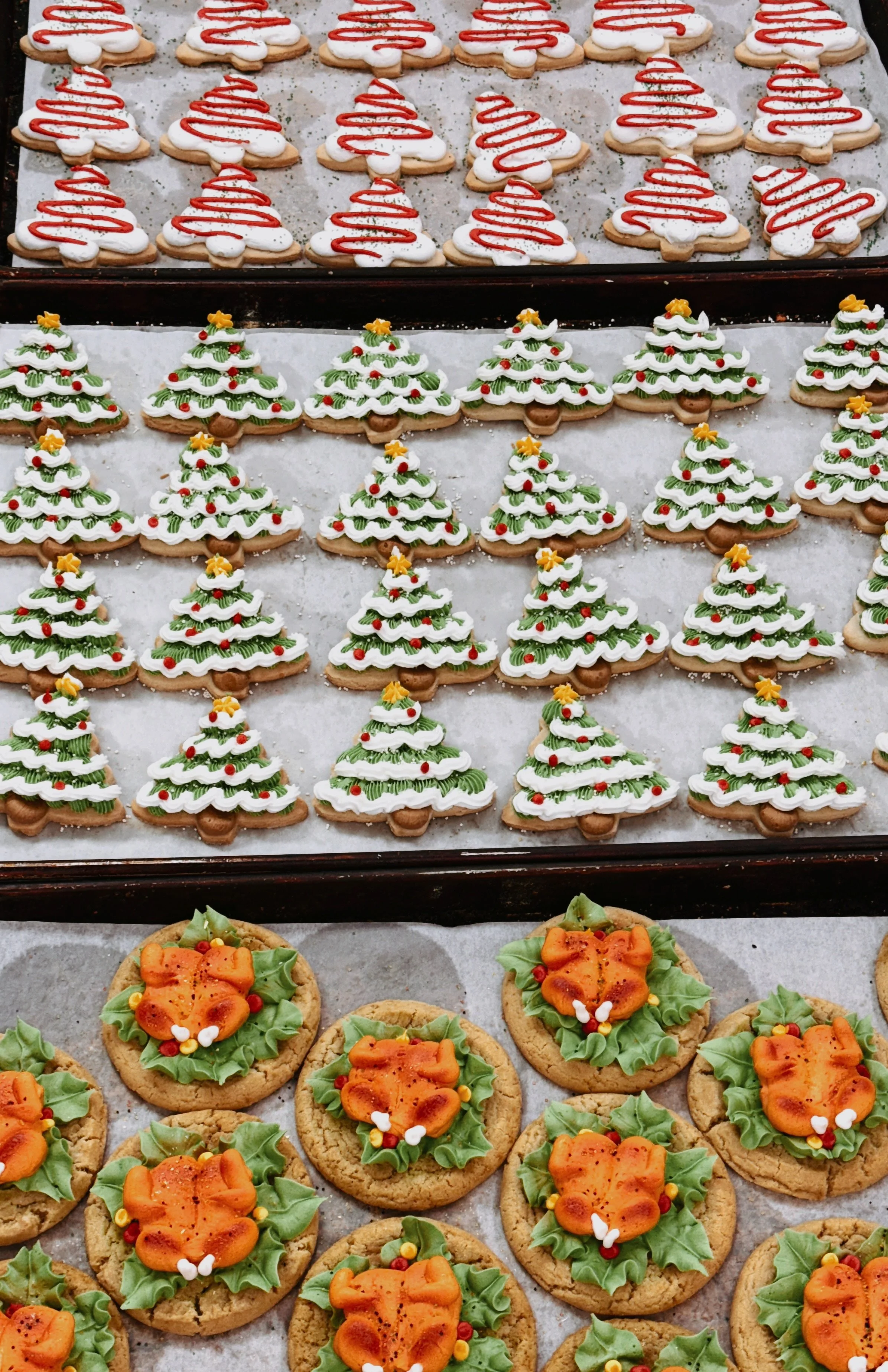 Assorted Christmas cookies arranged on trays, including tree-shaped cookies with white icing, Christmas tree designs with green icing and decorations, and cookies with orange flowers and green icing leaves.