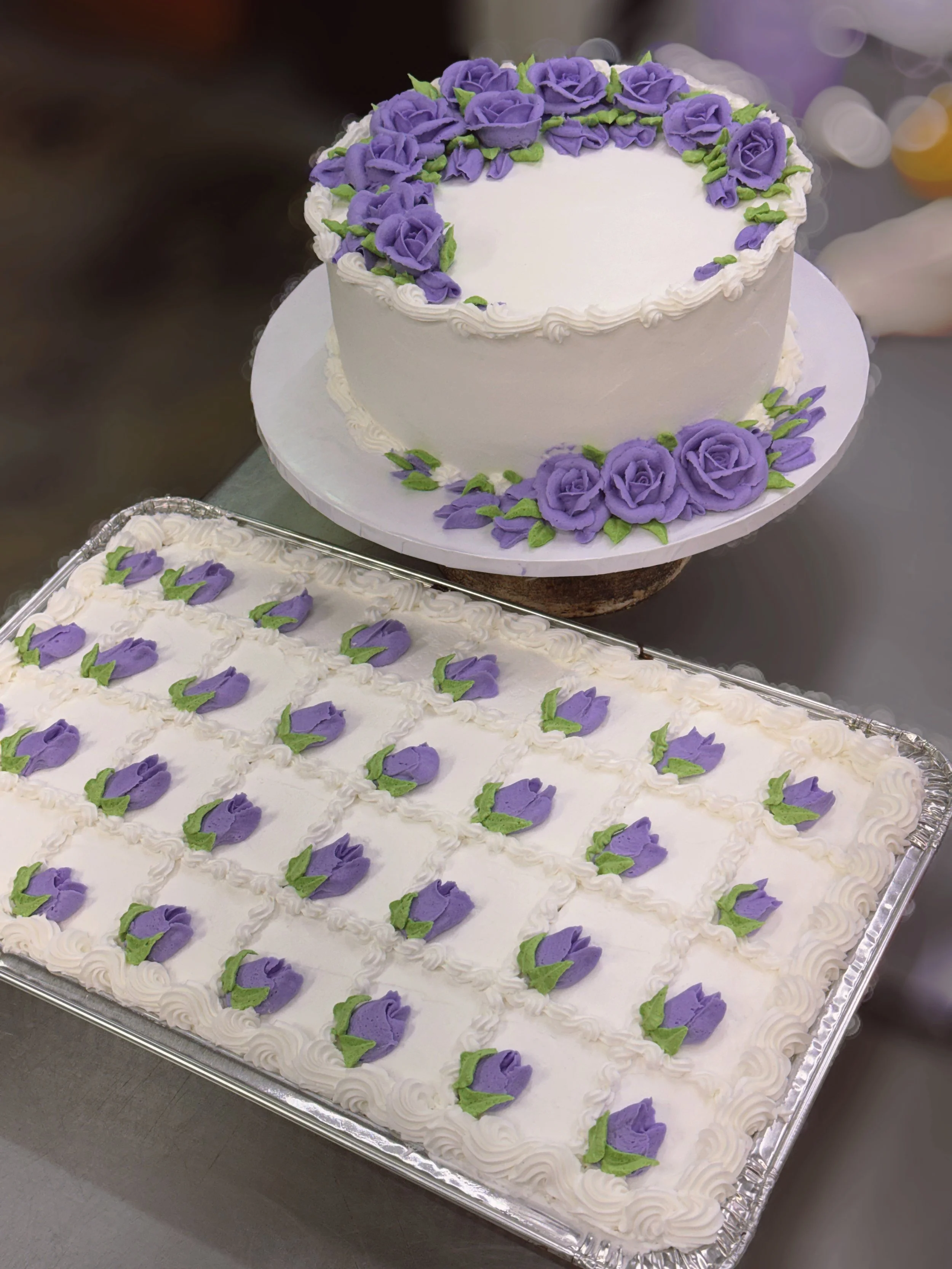 A white oval cake decorated with purple roses and green leaves, topped with a border of white frosting, on a silver tray.