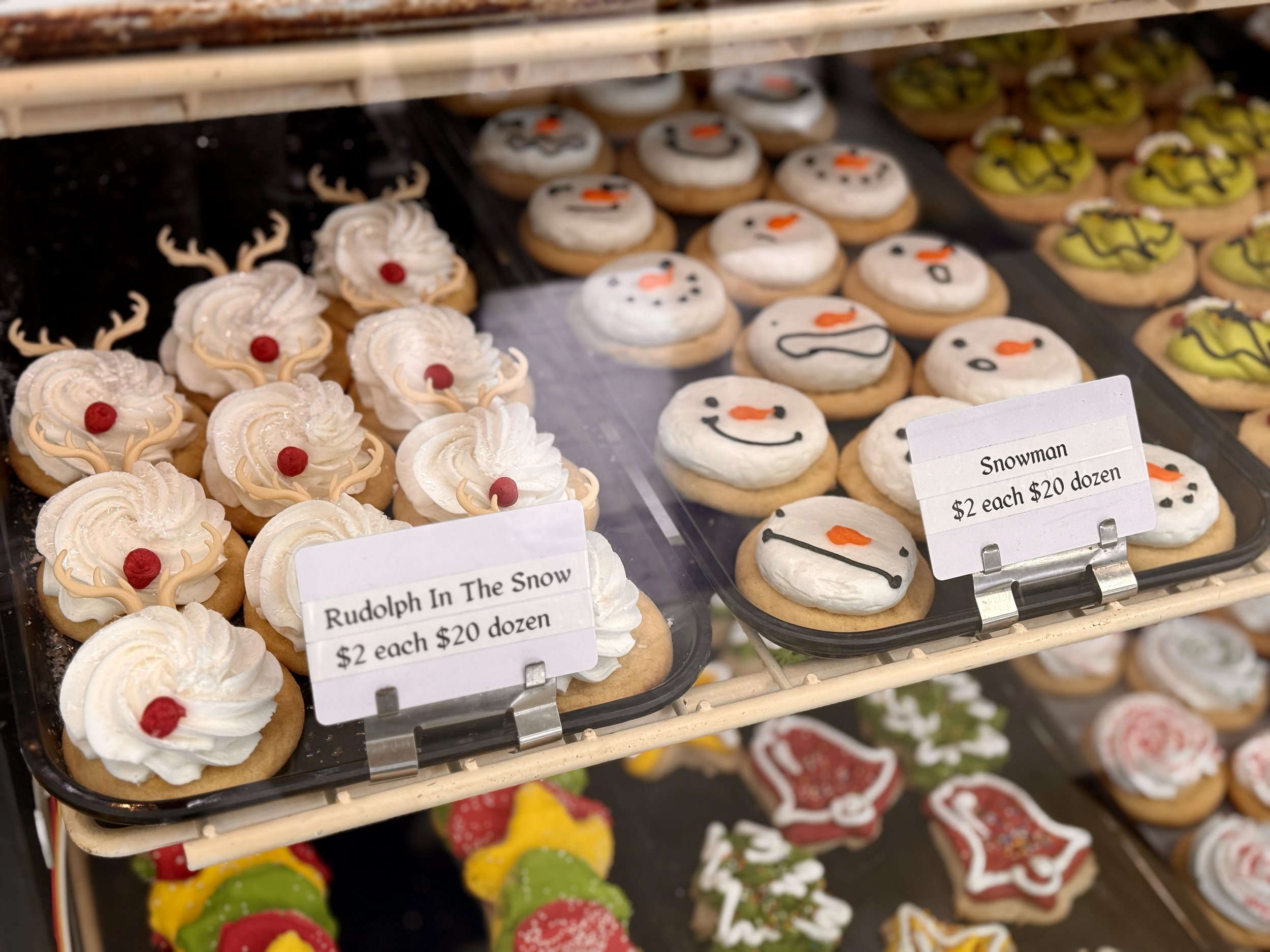 Display case with decorated Christmas cookies shaped like snowmen, reindeer, and various festive designs. Sign indicates prices for cookies.