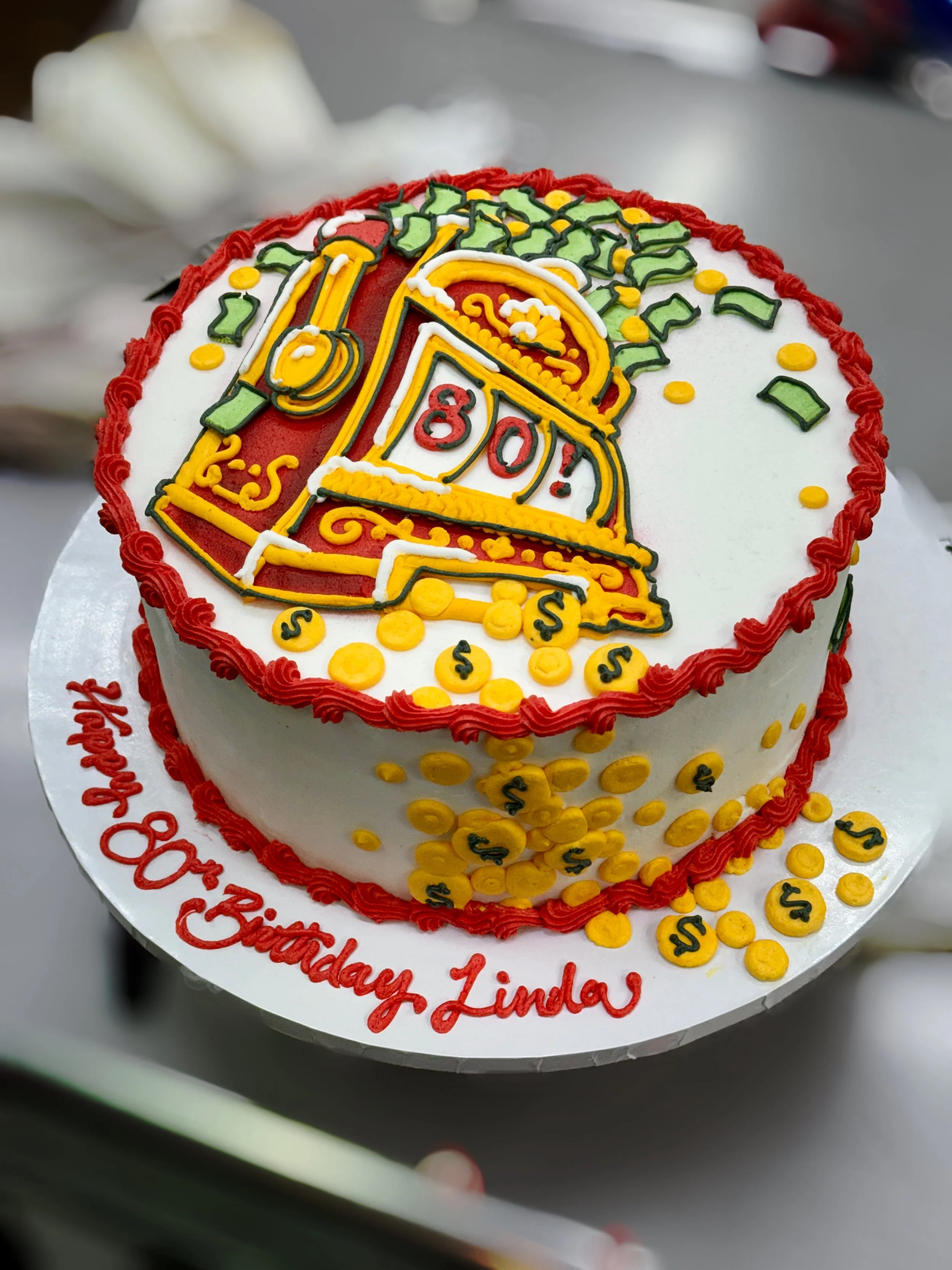 A birthday cake decorated with a money-related theme, featuring icing that depicts dollar signs, coins, and a cash register, with a message that reads 'Happy 80th Birthday Linda' in red icing.