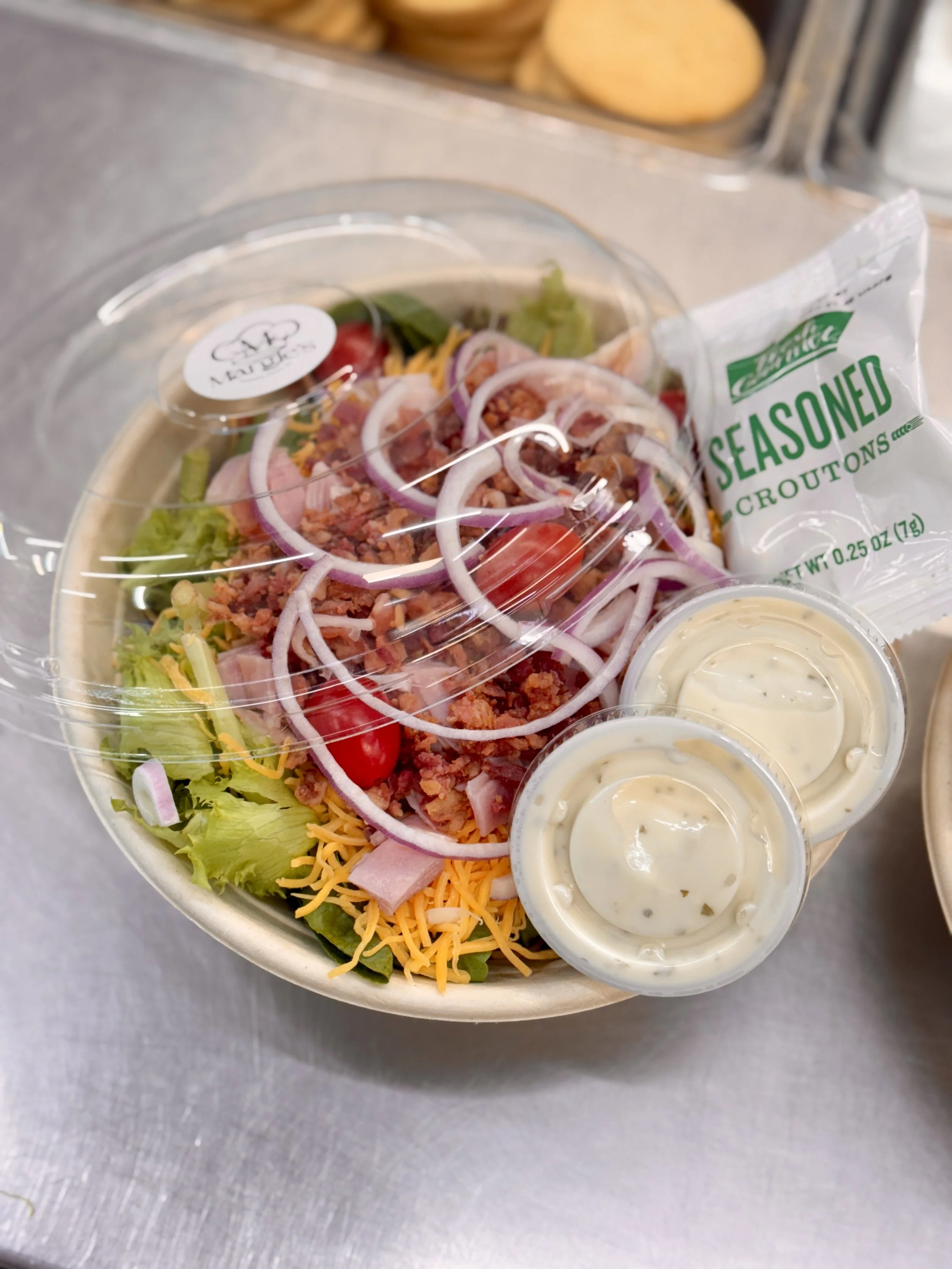 A plastic bowl of salad with shredded cheese, cherry tomatoes, chopped ham, sliced onions, and lettuce, with small containers of ranch dressing and a package of seasoned croutons nearby.
