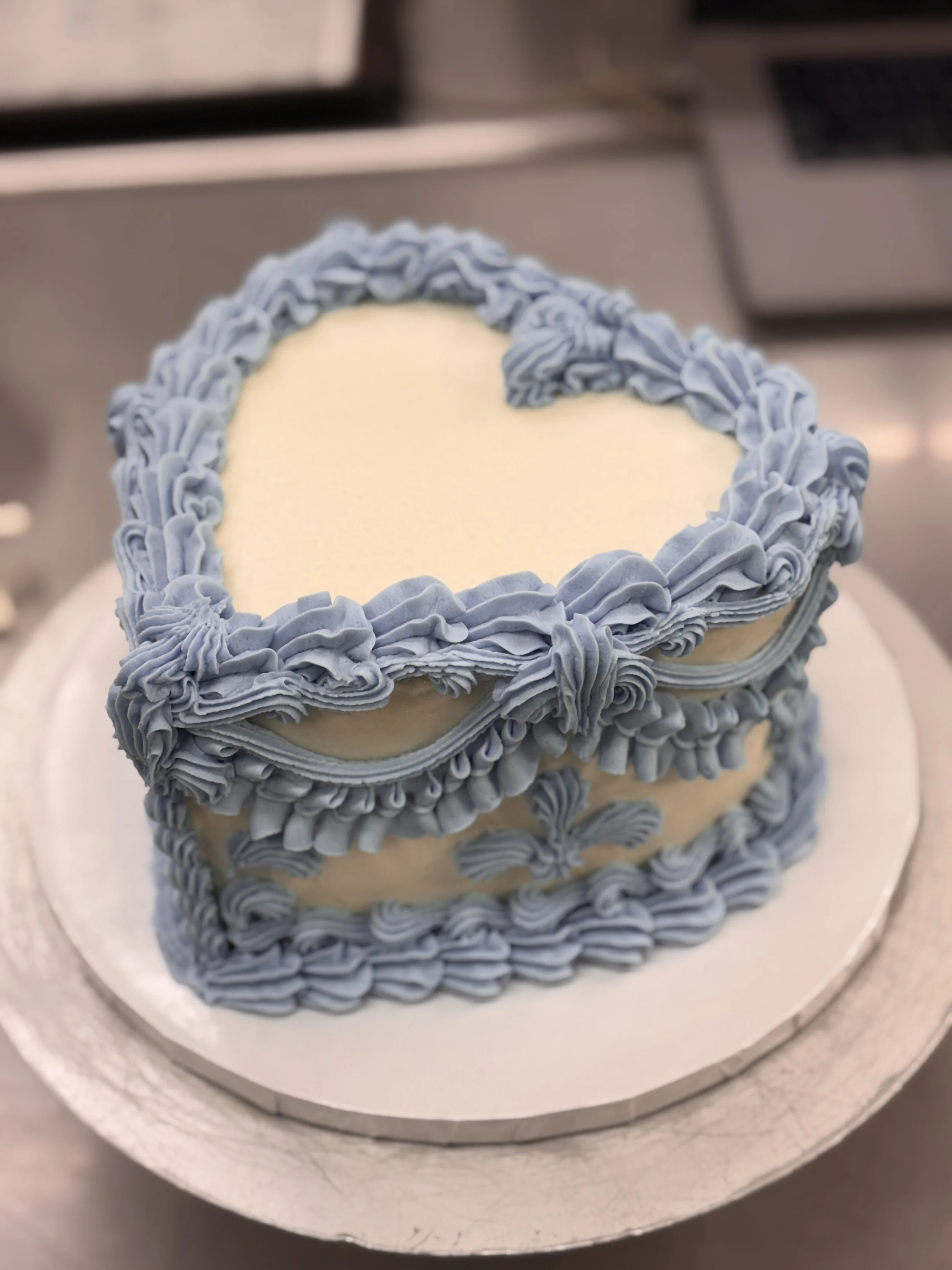 A heart-shaped cake decorated with ruffled light blue frosting and placed on a silver cake board.