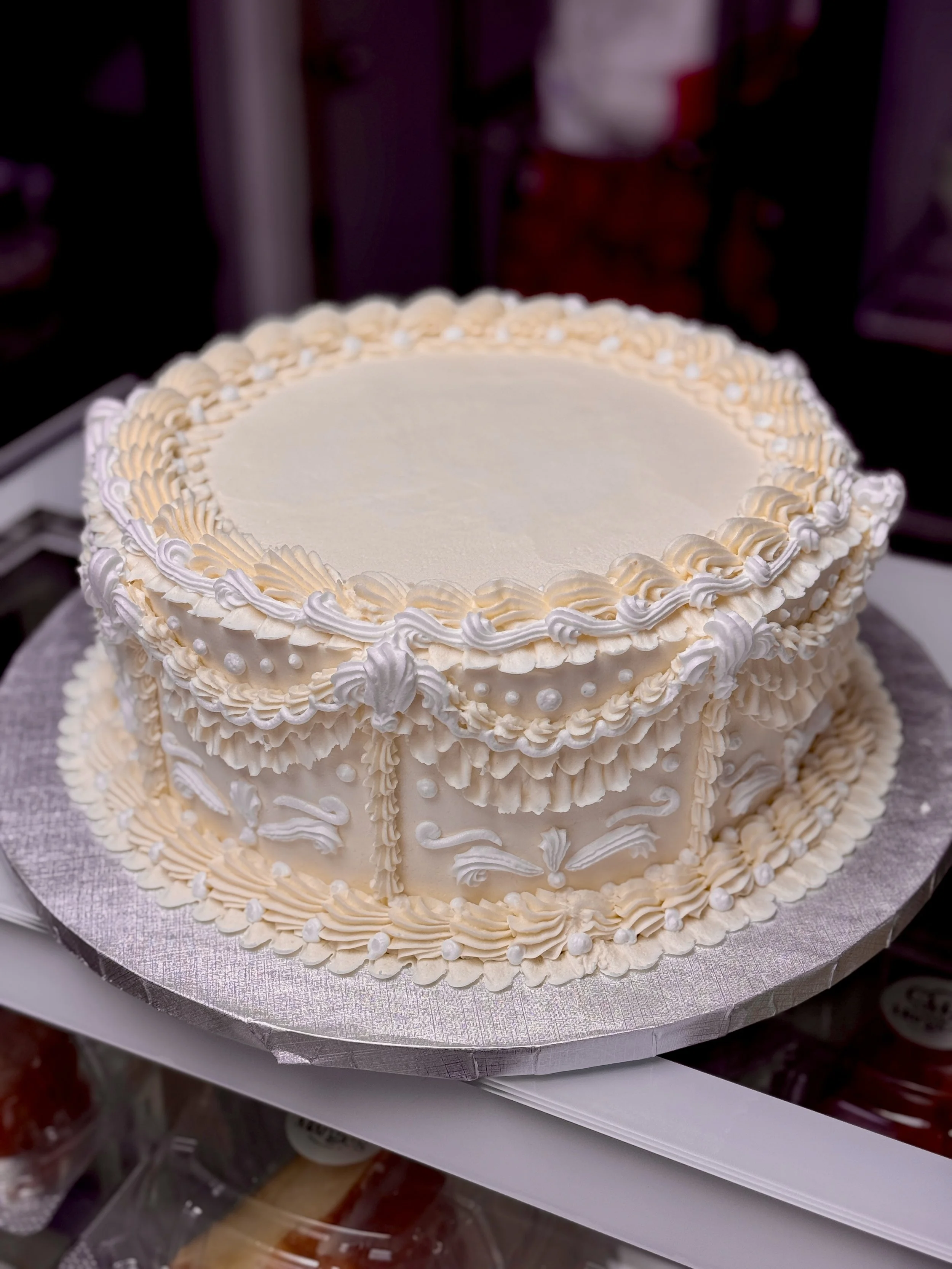 A round white cake decorated with intricate piping and small pearls on top, placed on a silver cake board.