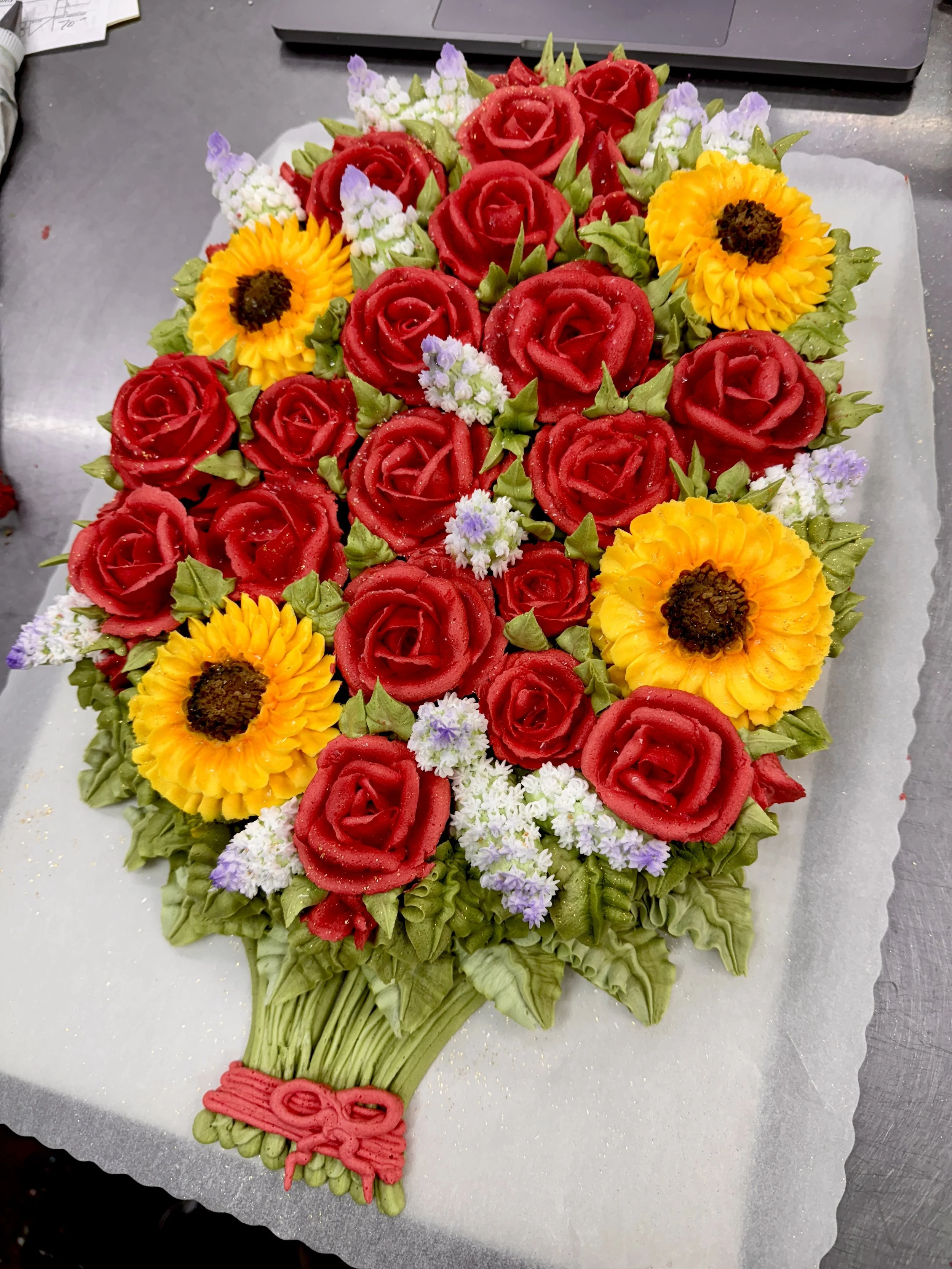 A floral bouquet arrangement with red roses, yellow sunflowers, small purple and white flowers, and green foliage.