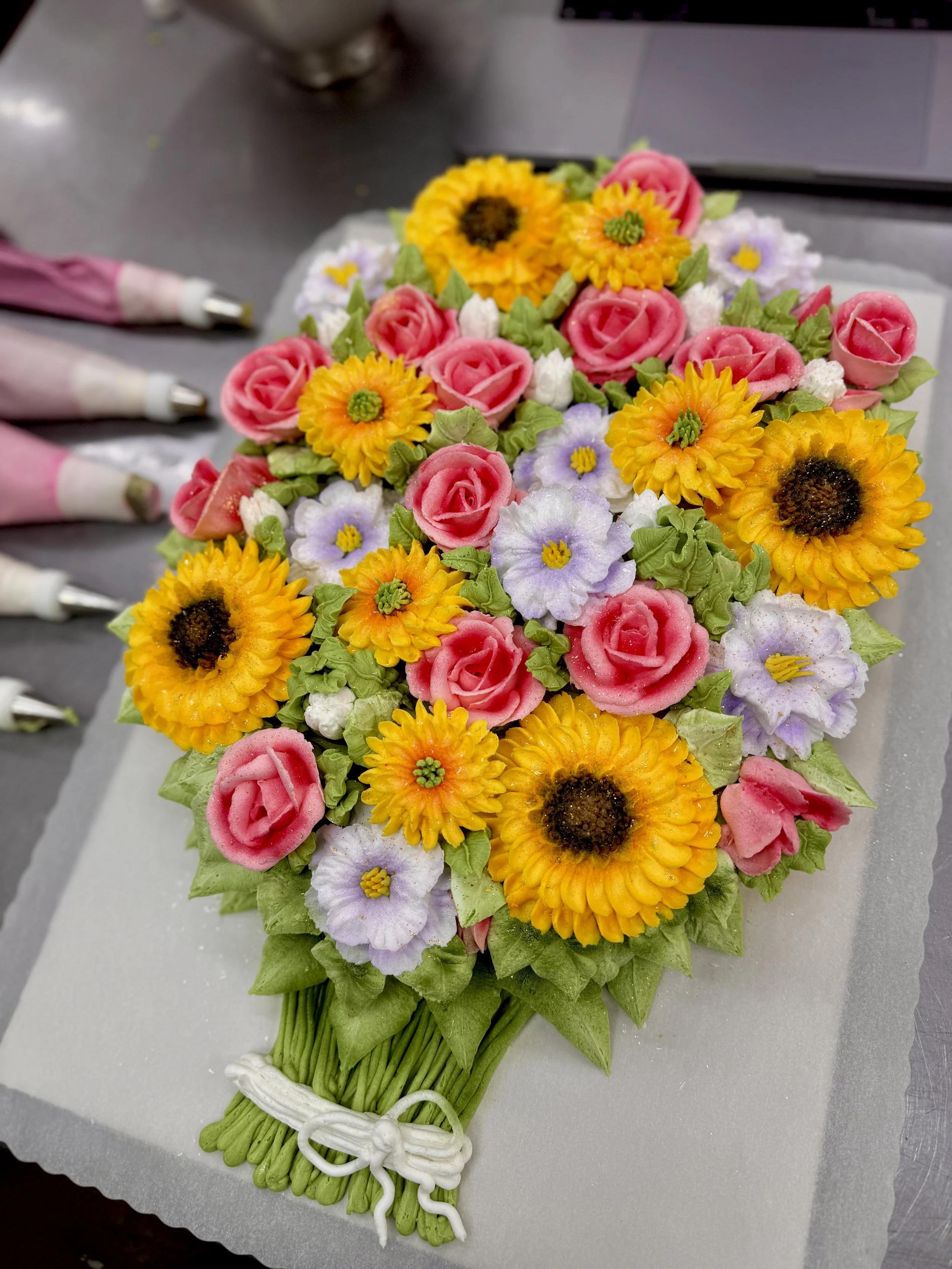 A beautiful bouquet of decorative flowers arranged with pink roses, yellow sunflowers, purple flowers, and green leaves, tied together with a white ribbon.