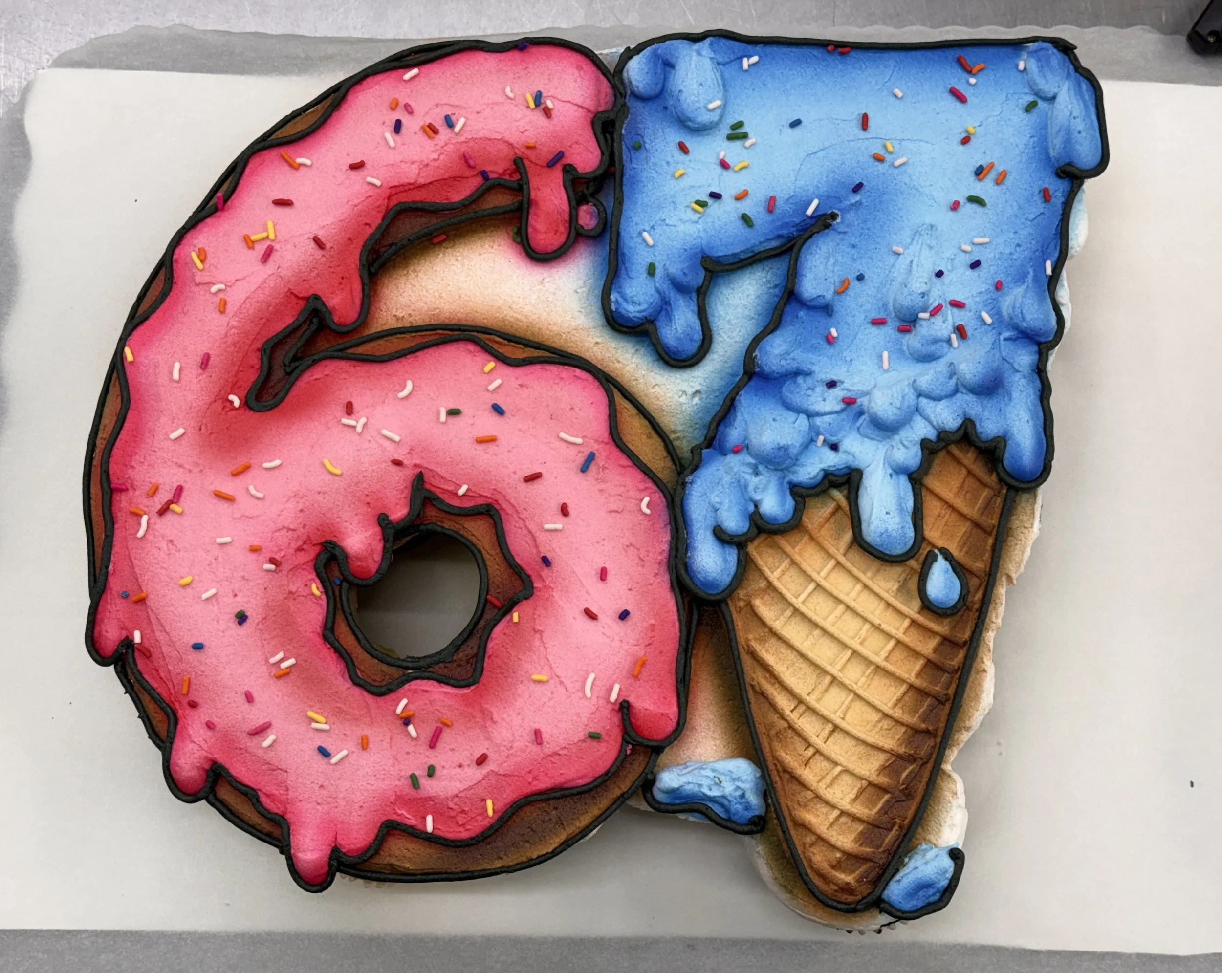 Decorated sugar cookies in the shape of the letter D, with pink and blue icing, colorful sprinkles, and outlined with black icing. The cookies are on a tray with a gray and white surface.