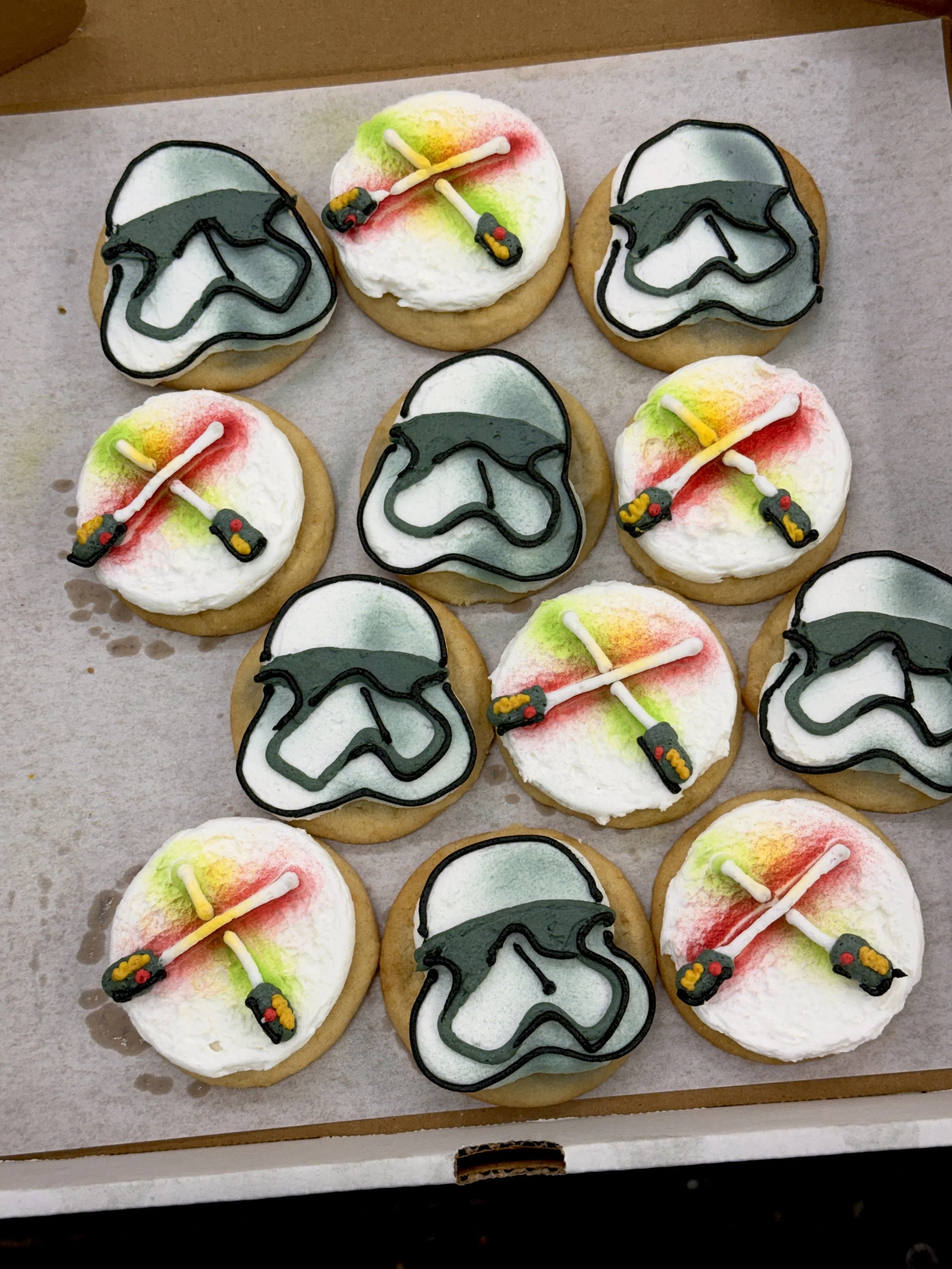 Decorated sugar cookies with white frosting, black and green icing outlines, and colorful firework and lighter motifs on top, set on parchment paper in a white box.